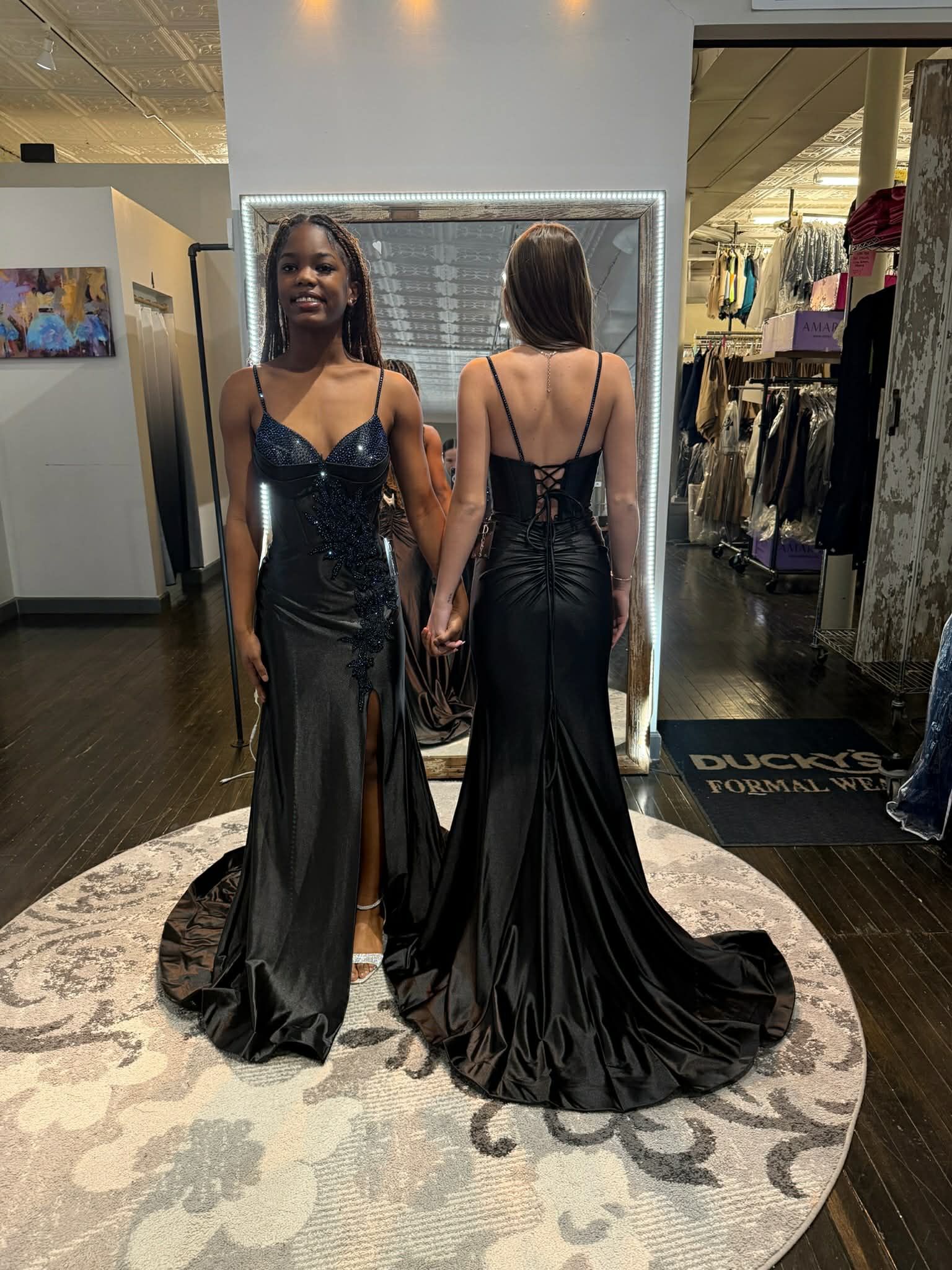 A person in a black, floor-length, shimmering gown with a high leg slit and lace-up back stands before a large mirror.
