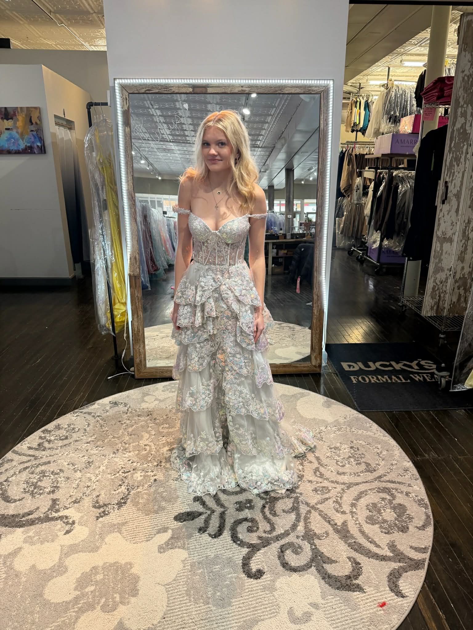 A person wearing a tiered, off-the-shoulder gown stands in front of a mirror in a bridal boutique.