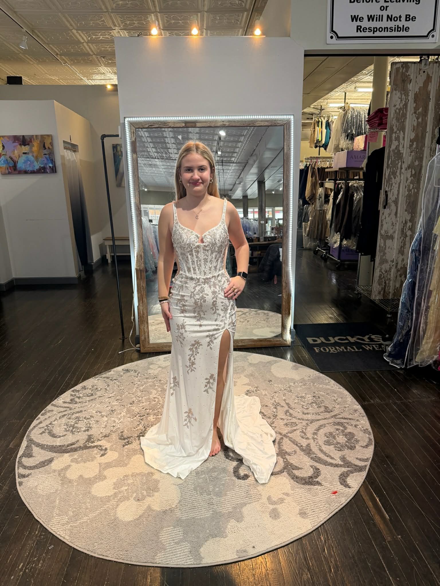 A person stands on a round rug in a store, smiling while wearing a strapless, white gown with pink floral patterns.