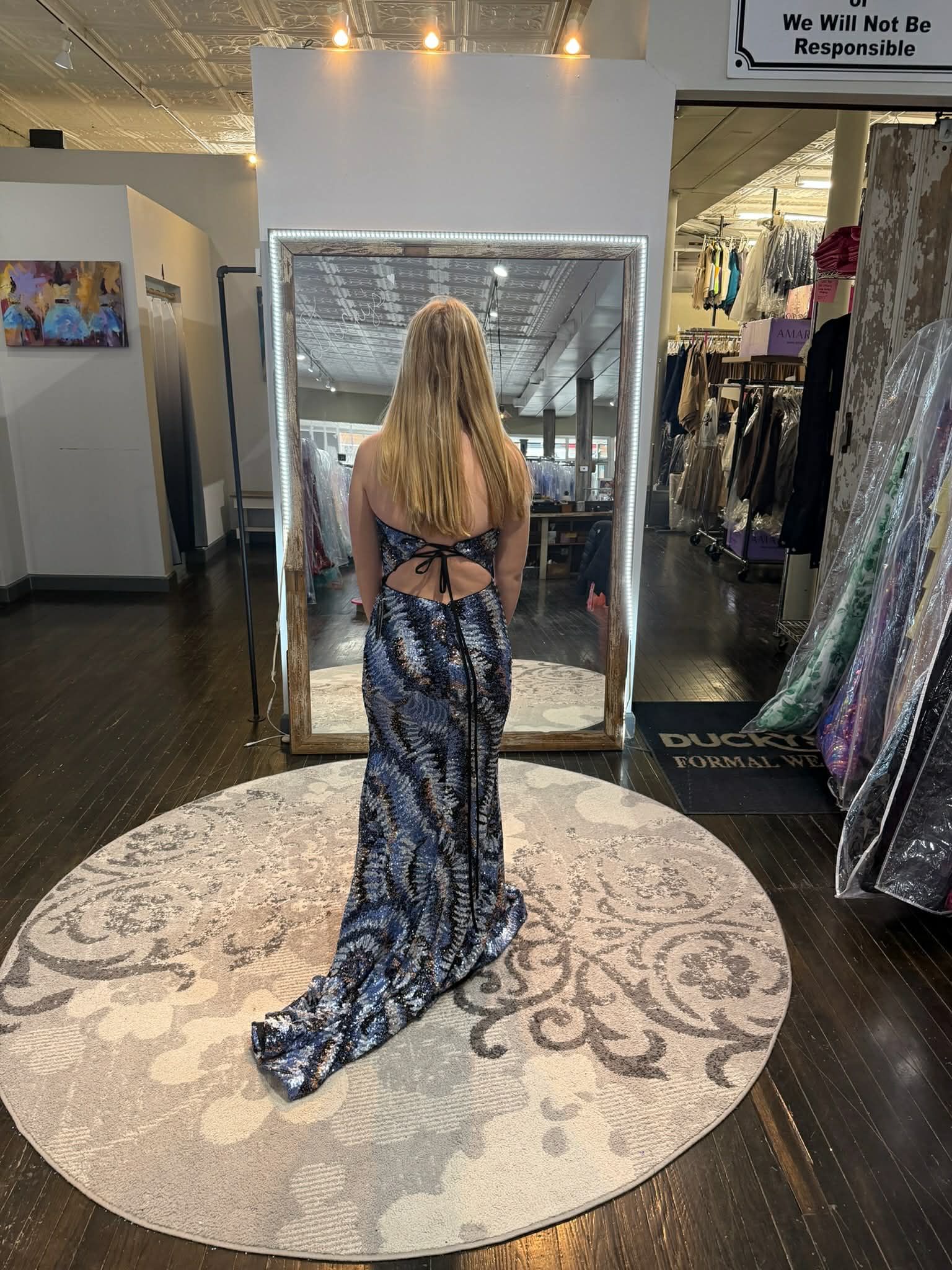 Seven people stand in a clothing boutique wearing colorful, floor-length formal gowns, arranged in a row on a round rug.