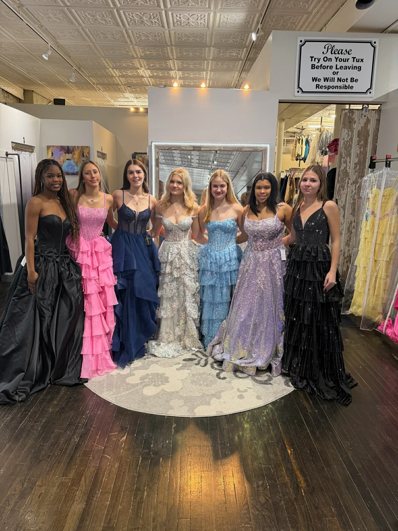 A smiling person stands on a circular rug in a boutique, wearing a light blue, sequined floor-length formal gown.