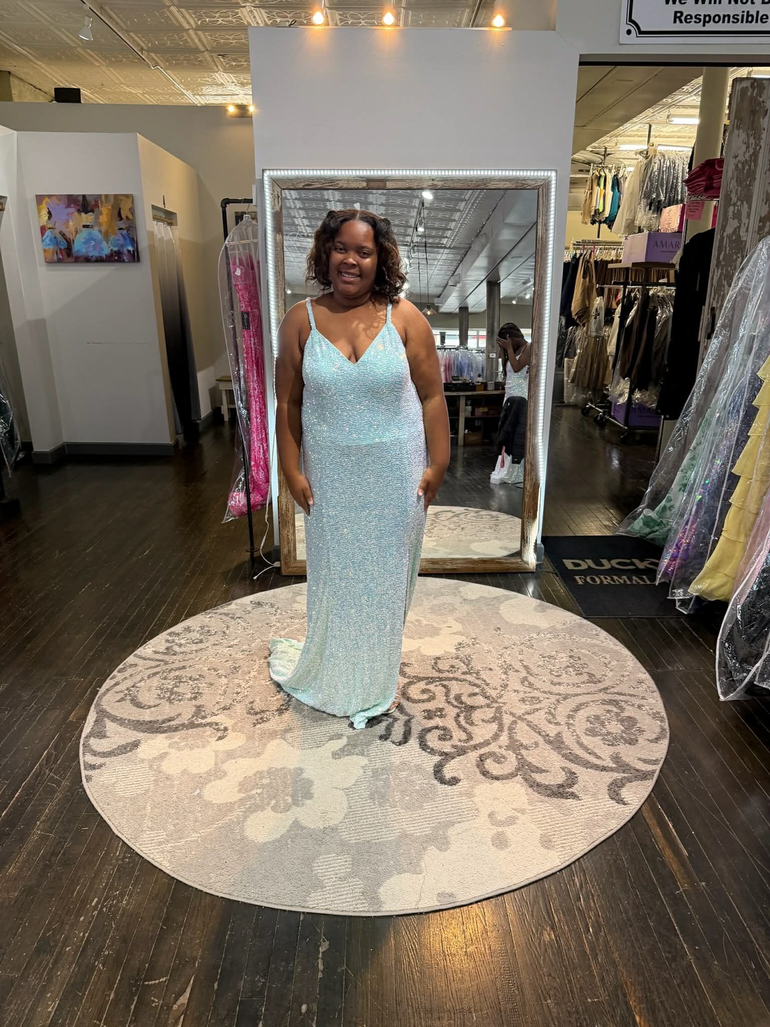 A person wearing a tiered light blue gown stands on a patterned rug in front of a large mirror in a clothing store.