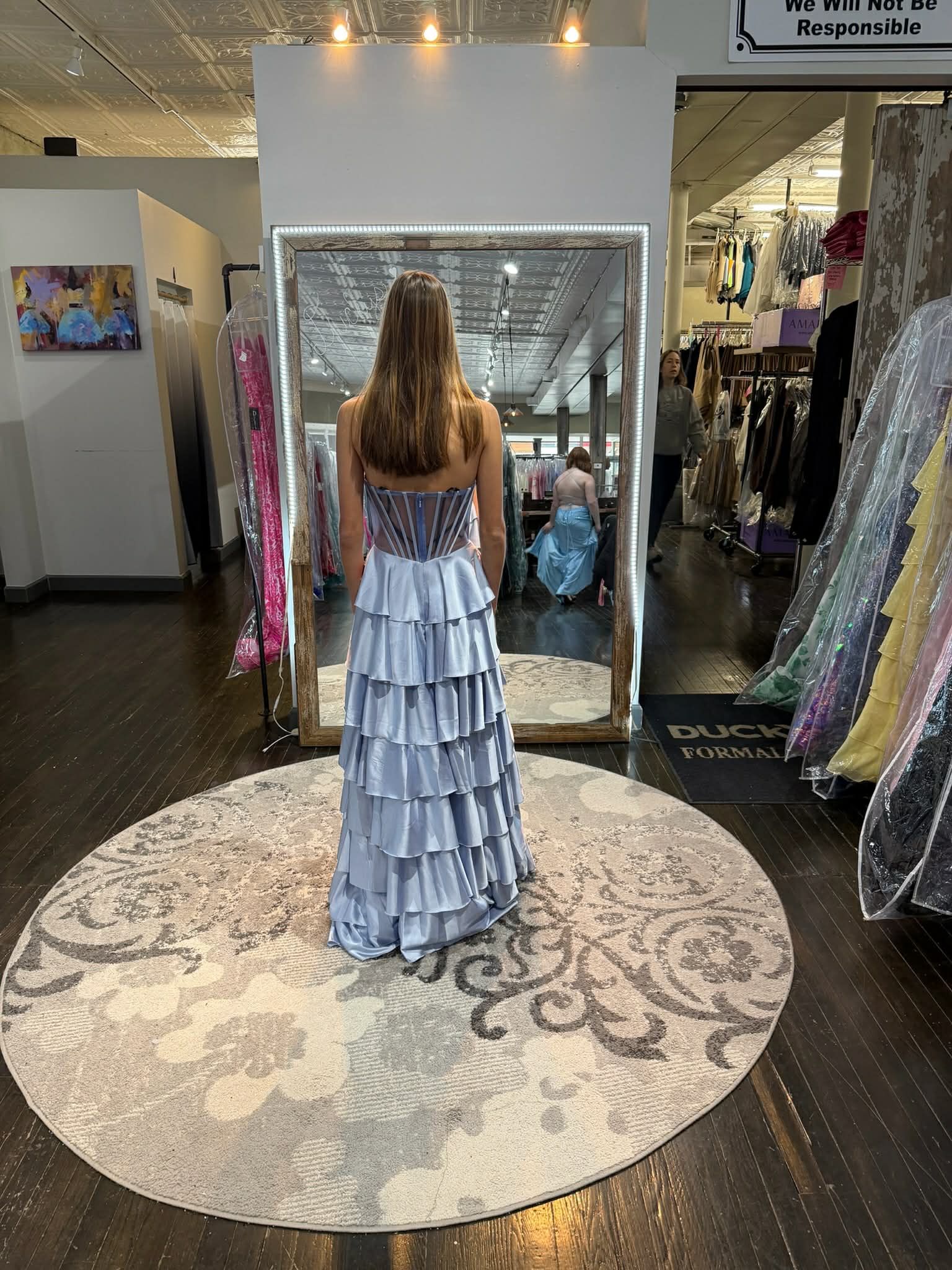 A person stands on a round rug in a clothing store, wearing a sparkling red gown while facing a large mirror.