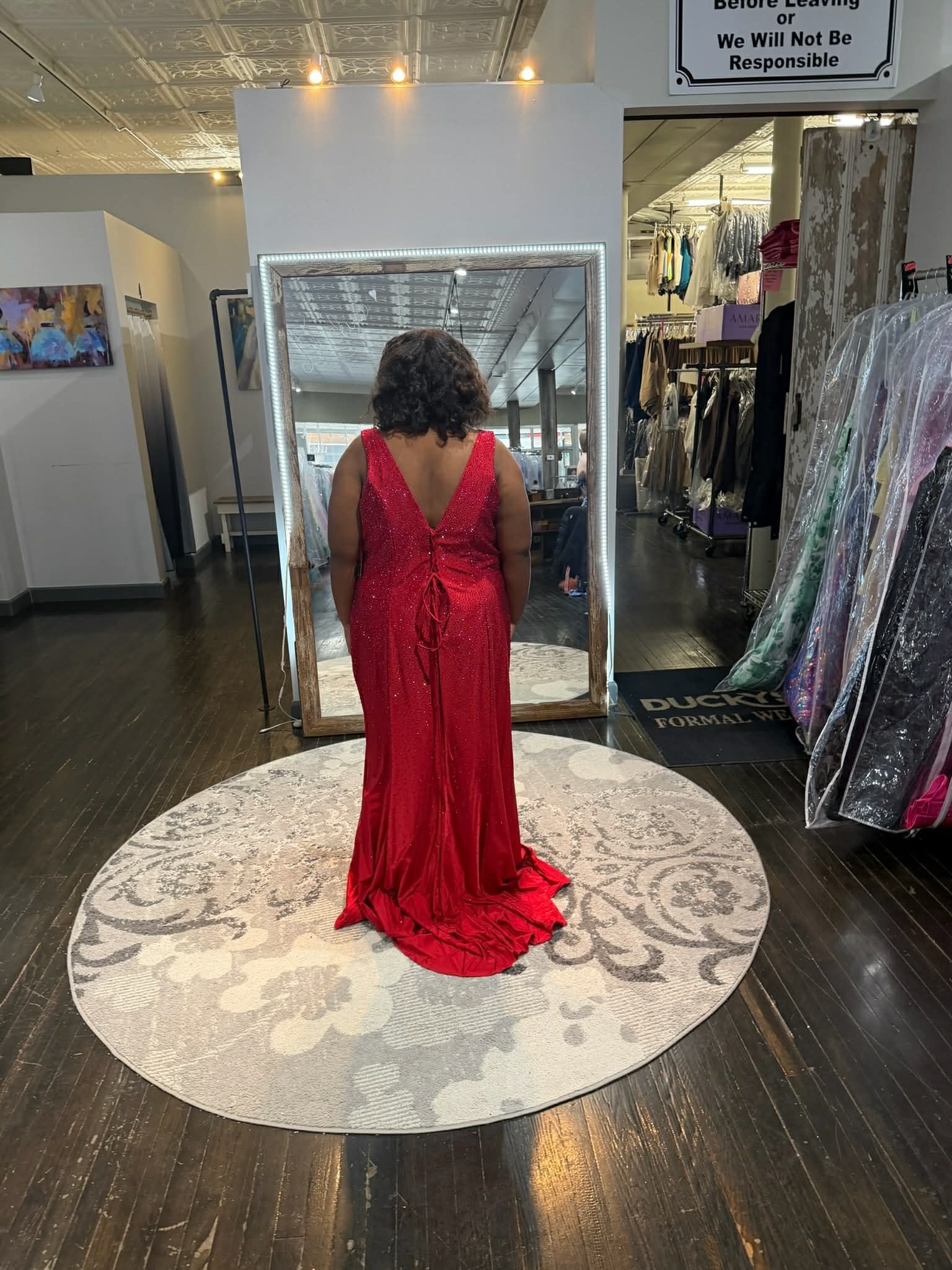 A person with red hair stands on a circular rug in a boutique, wearing a lavender floor-length gown with ruffled layers.