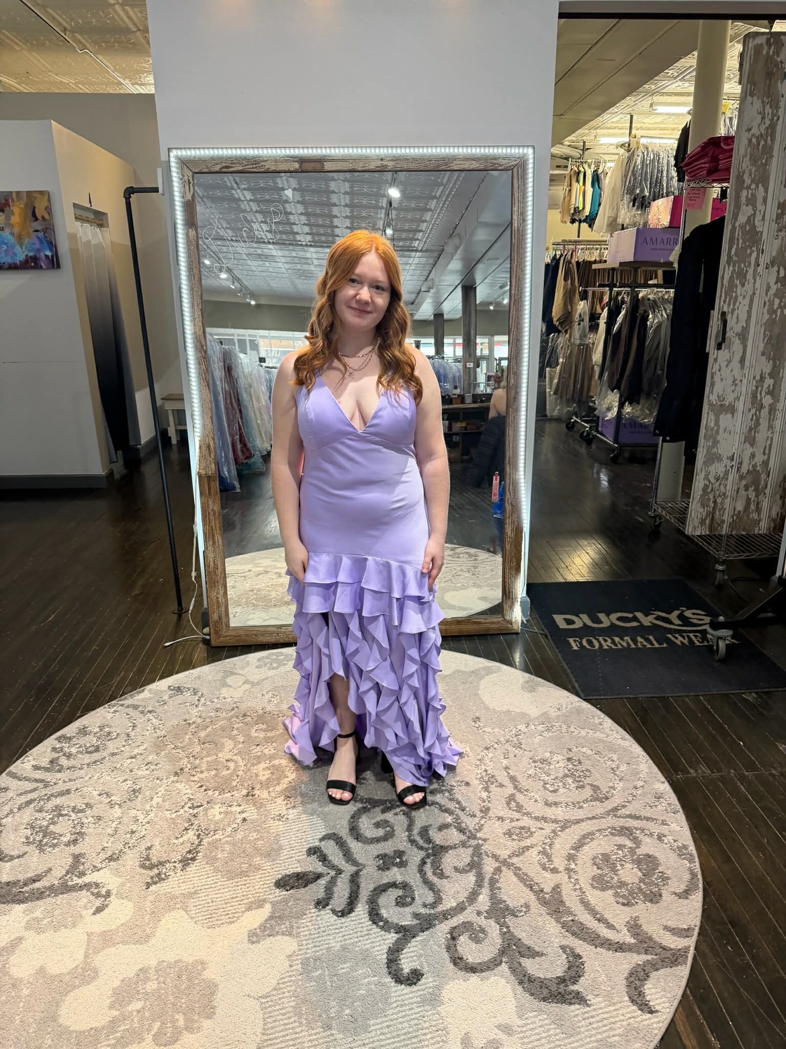 A person stands on a circular platform in a clothing store, wearing a shimmering, dark green, floor-length gown with a slit.