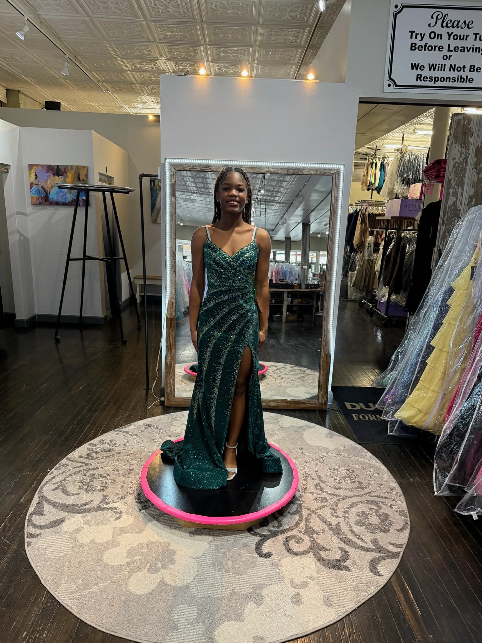 A person stands on a patterned rug in a clothing boutique, facing a mirror while wearing a vibrant magenta floor-length gown.
