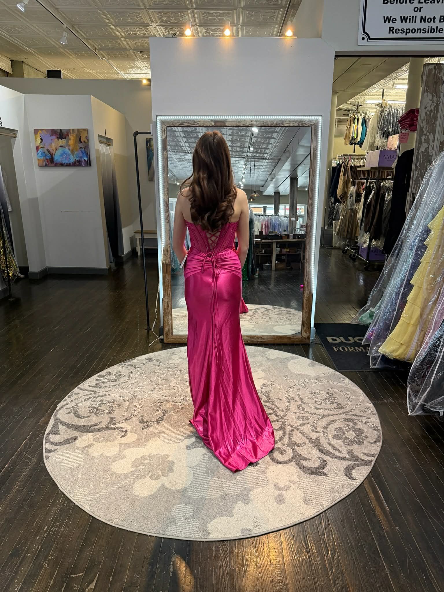 A person stands on a circular rug in a boutique, wearing a bright pink, floor-length gown with a lace bodice and a slit.