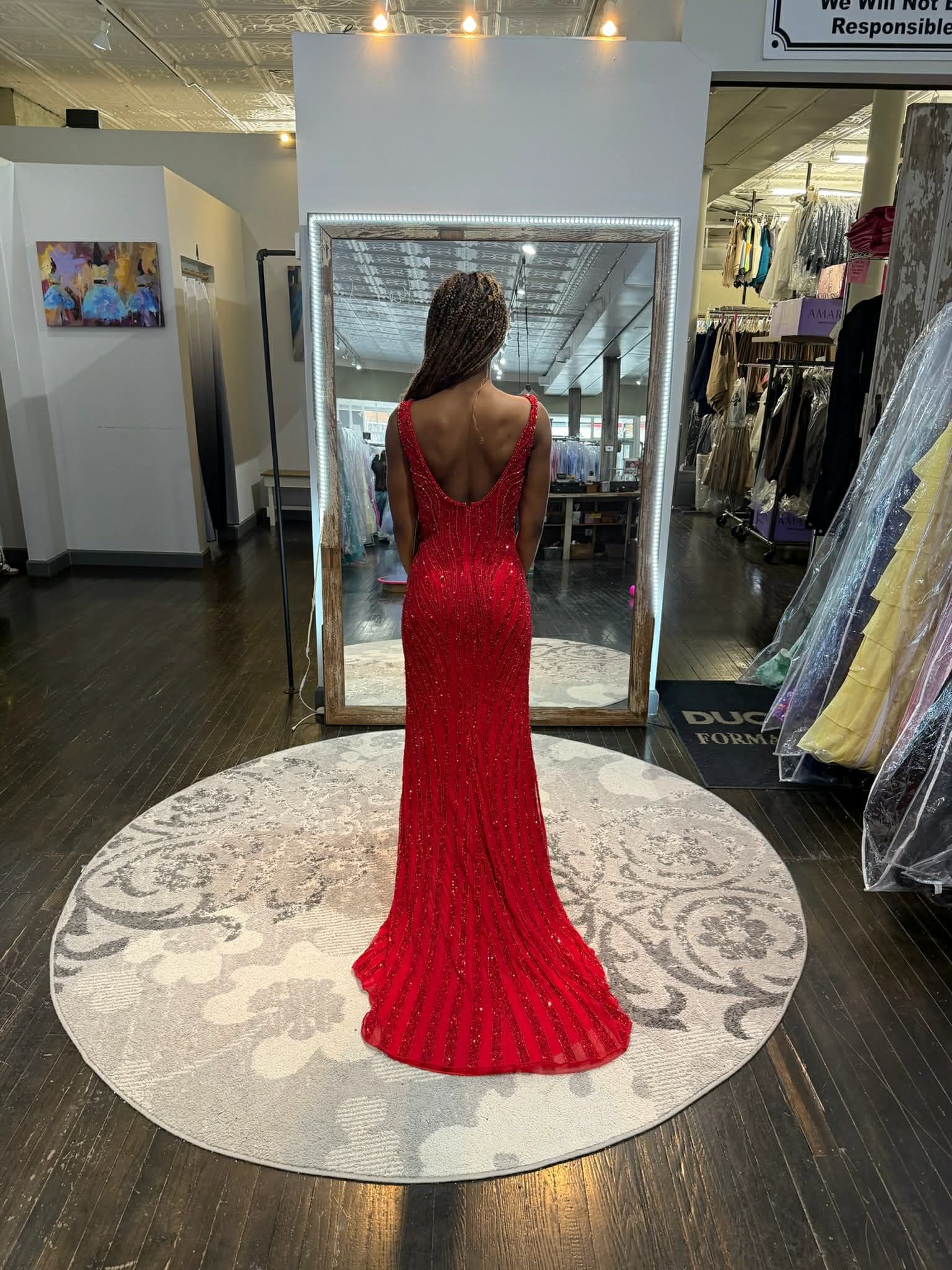 A person stands on a circular rug in a boutique, viewing their back in a large mirror while wearing a red formal gown.