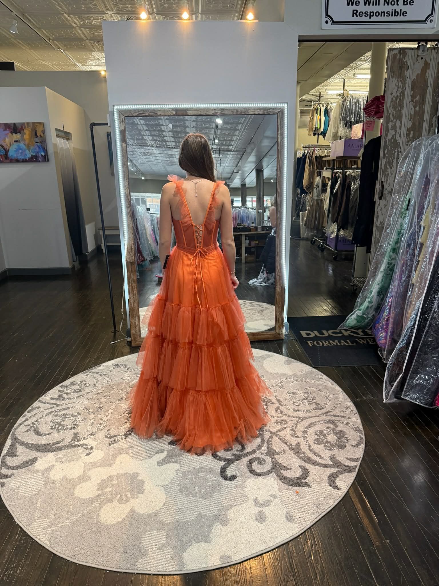 A person stands in a clothing store mirror wearing a floor-length, shimmering gold gown with a lace-up back and train.