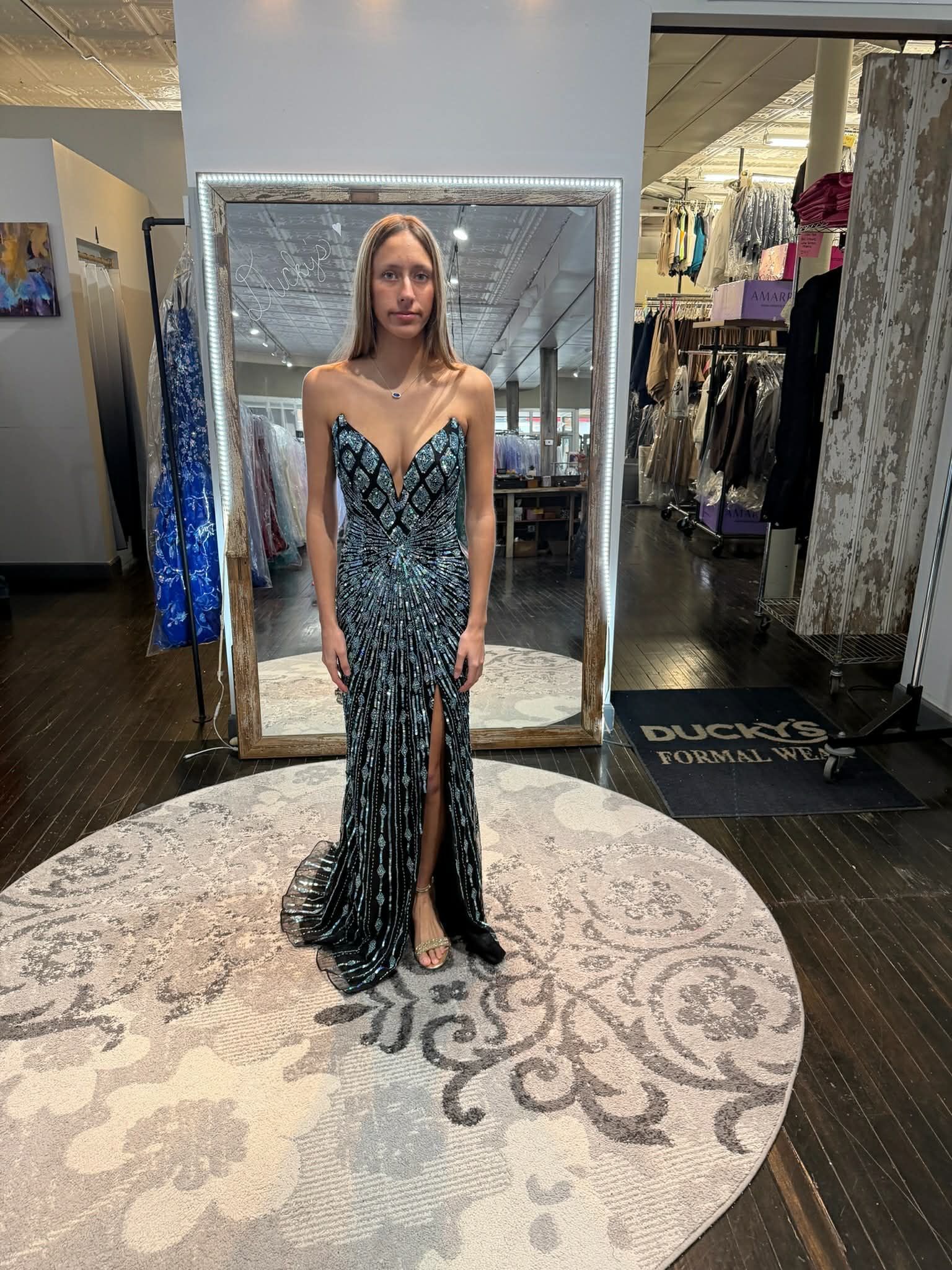 A person stands in a clothing store wearing a lavender, floor-length gown with a beaded bodice and a side leg slit.