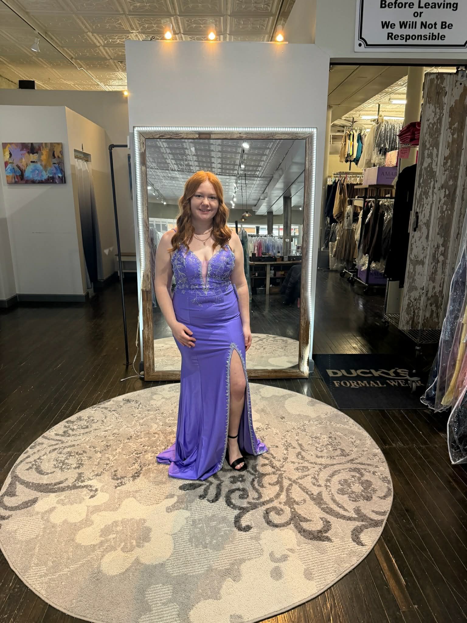 A person stands on a circular rug in a boutique, wearing a long, backless, purple-and-black sequined gown with a train.