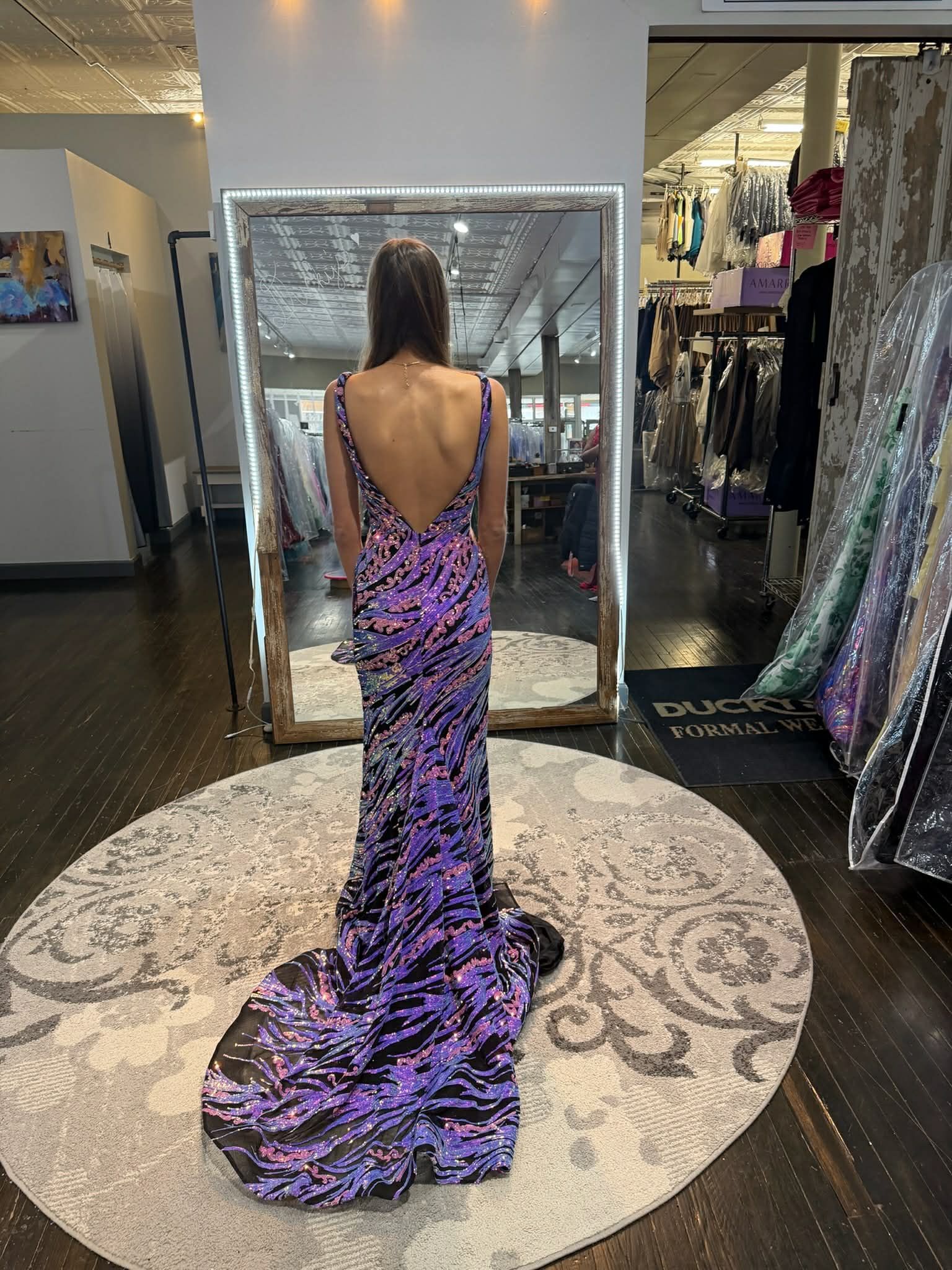 A person with blonde hair wearing a floor-length, light blue sequined gown stands on a circular rug in a boutique.