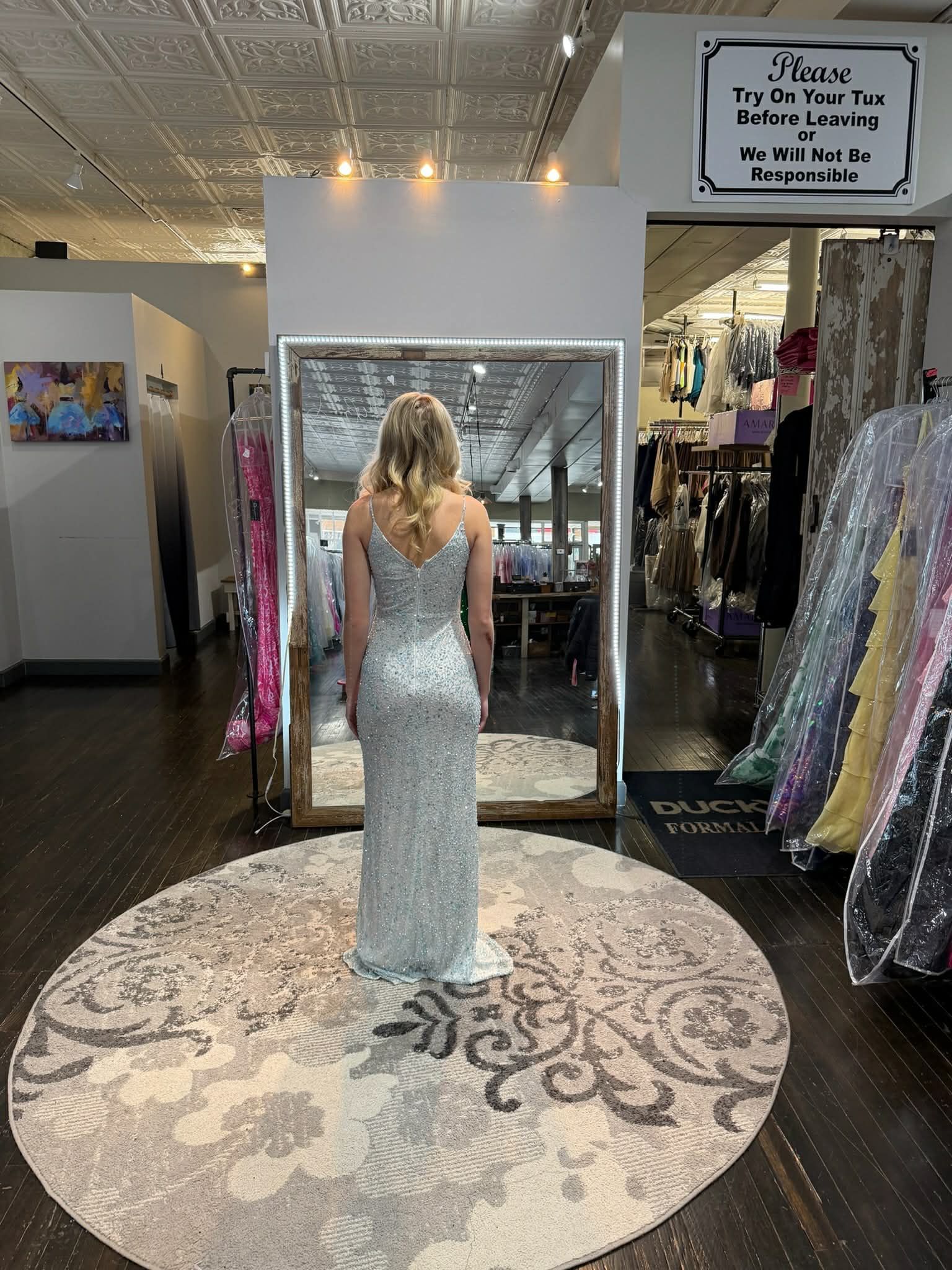 A person stands in a clothing store, facing a mirror while wearing a floor-length, bright pink gown with a lace bodice.