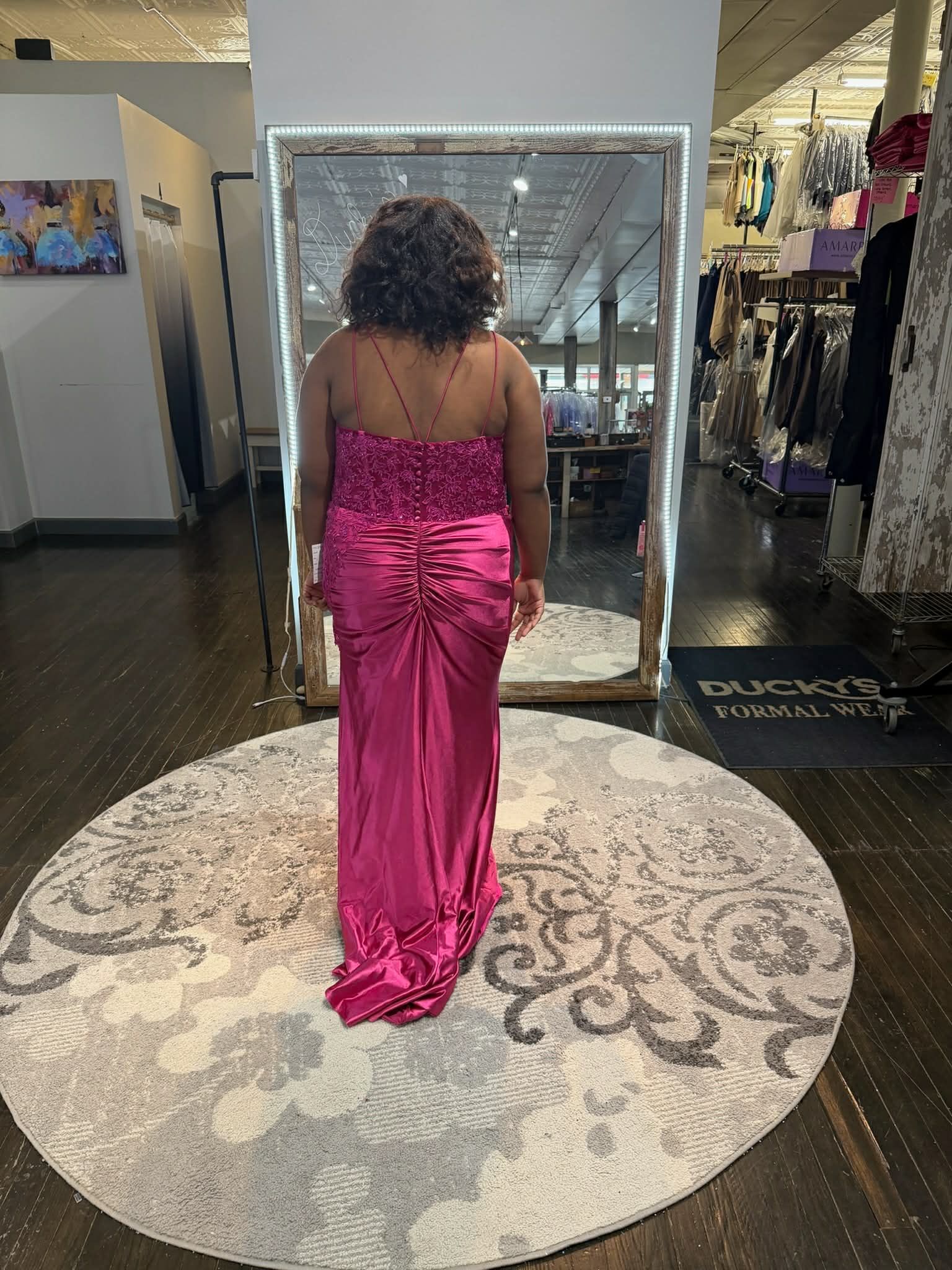 A person with long reddish hair smiling while wearing a sparkly red floor-length gown in a clothing store mirror.