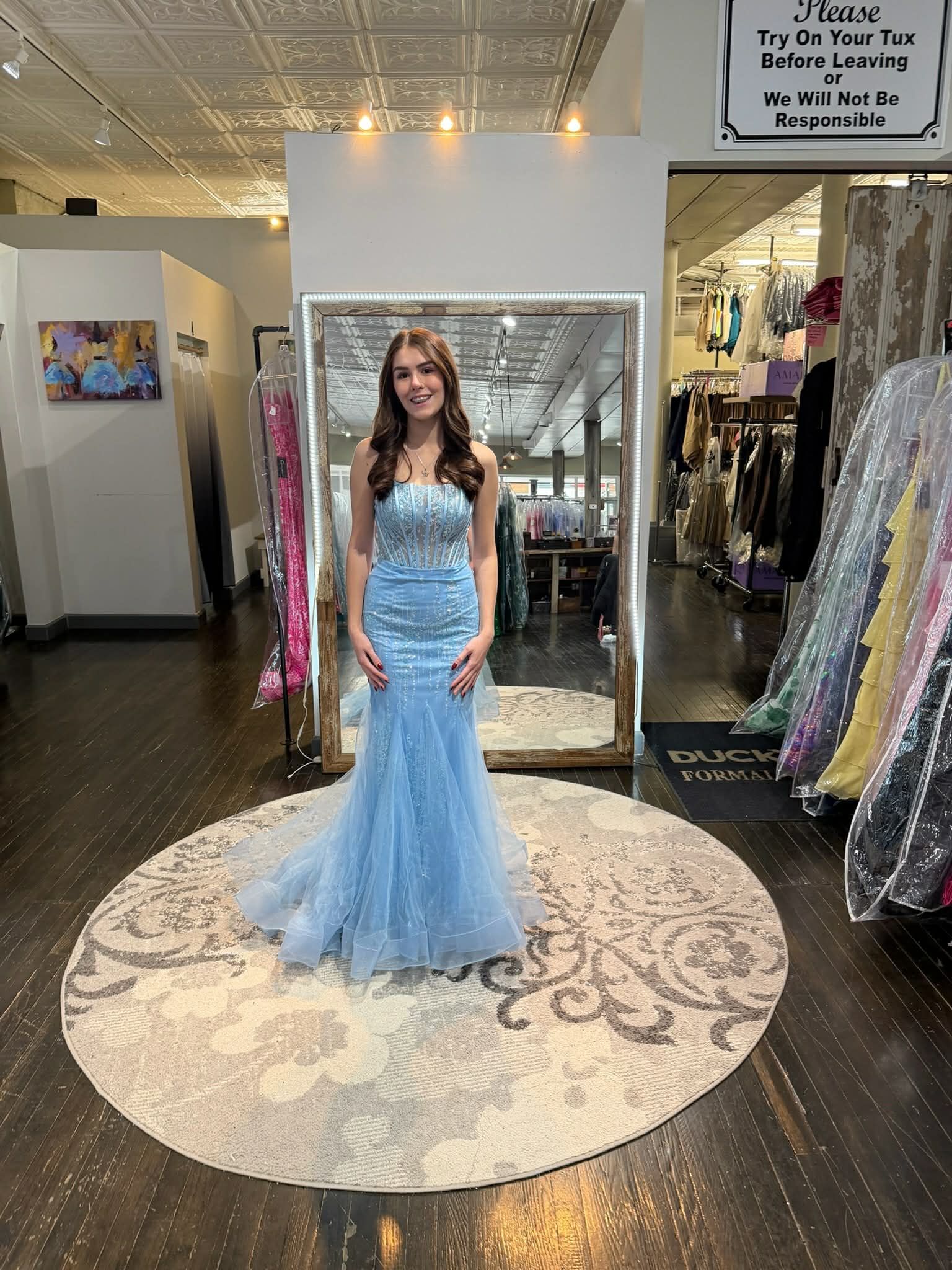 A person wearing a floral print gown with a train stands on a circular rug in front of a mirror in a clothing store.