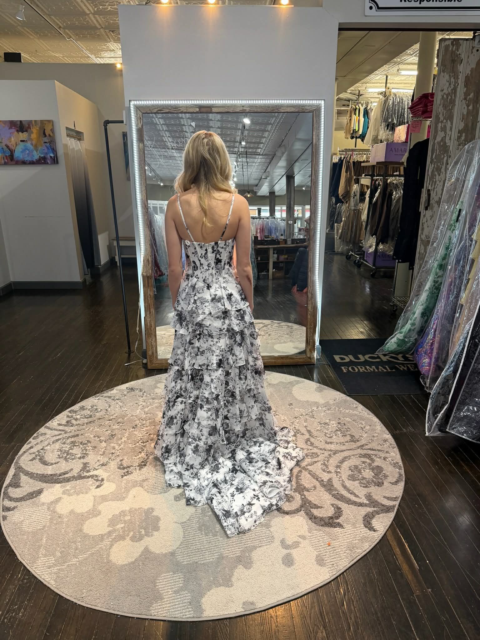 A person stands facing a mirror wearing a tiered, silver sequined off-the-shoulder gown in a retail clothing store.