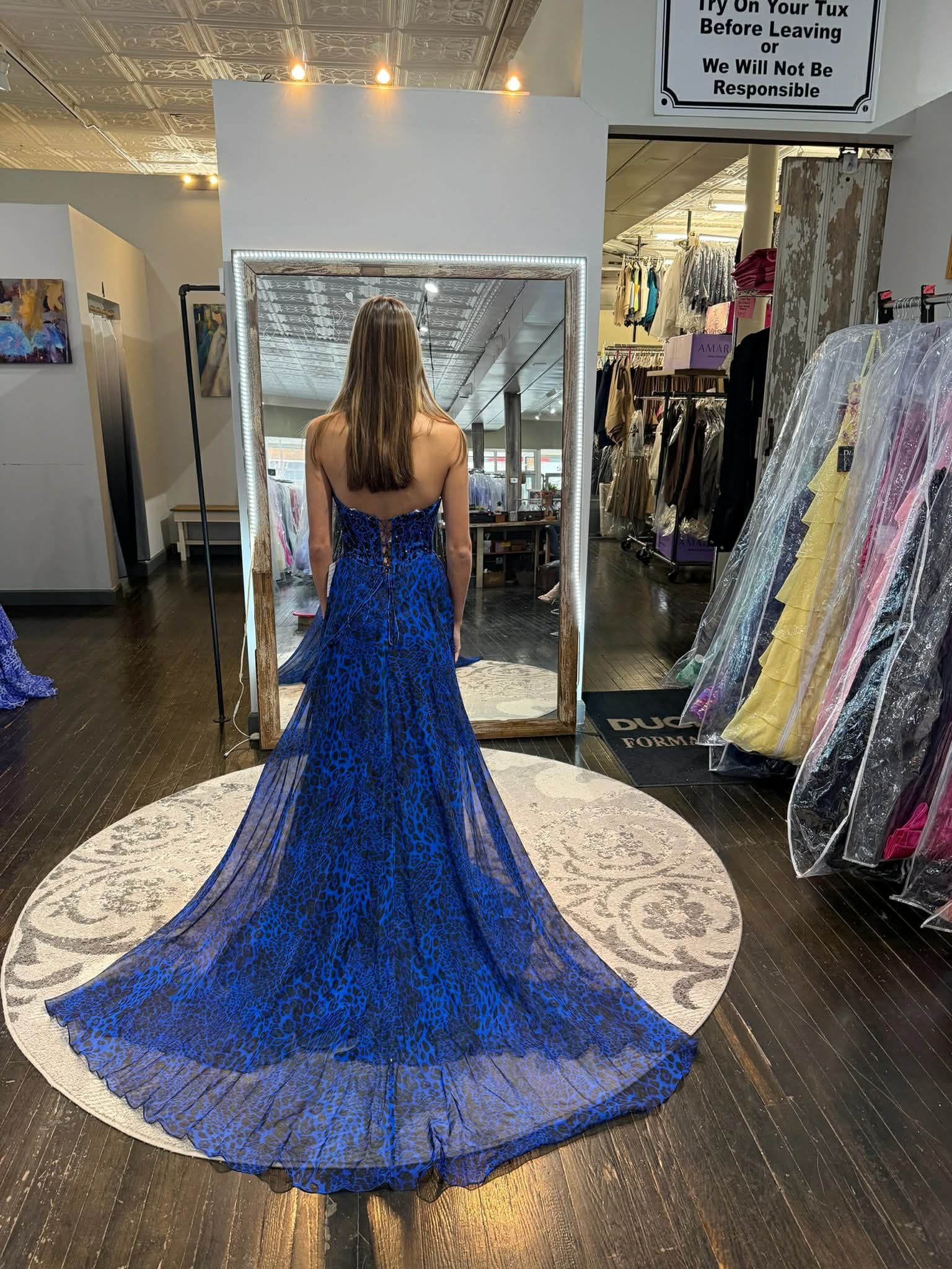 A person stands in a dress shop, facing a mirror and wearing a floor-length, light-colored sequined evening gown.