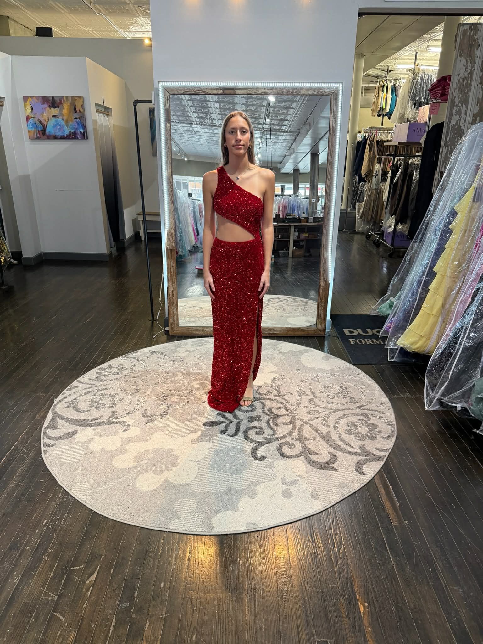 A person stands on a round rug in a boutique, wearing a floor-length emerald green sequined dress with a side slit.