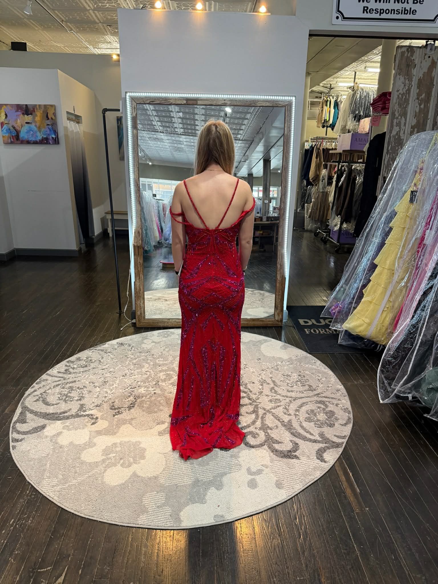 A person standing on a round rug in a clothing store, facing a mirror while wearing a magenta floor-length gown.