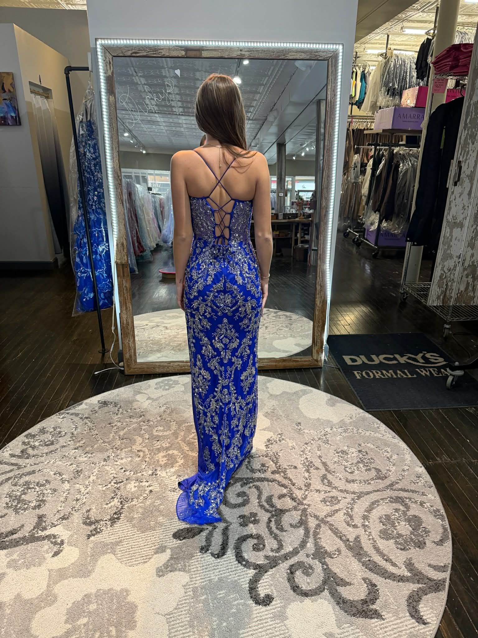 A person stands in a dress shop wearing a floor-length, shimmering gold sequined gown with a lace-up back design.