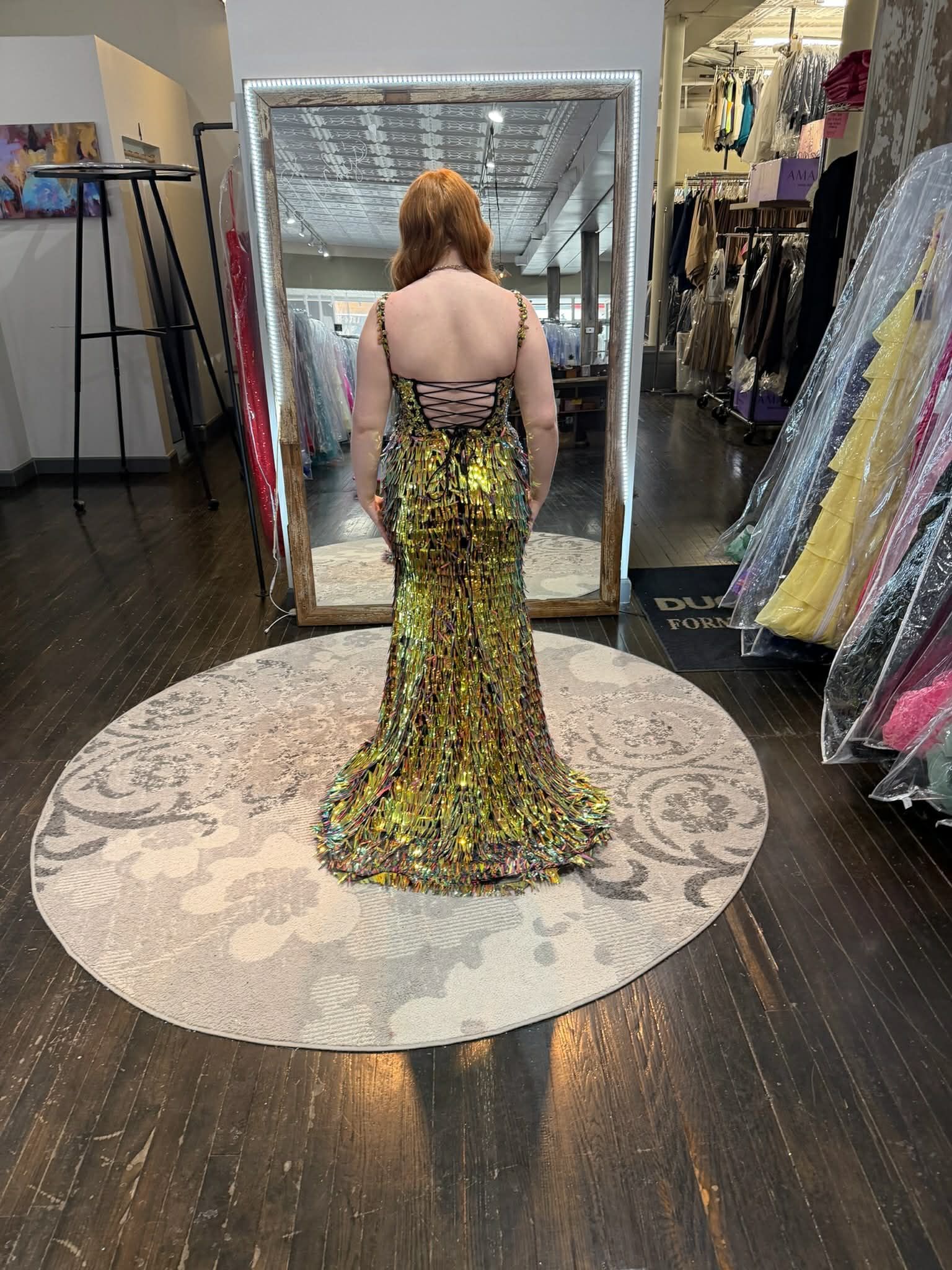 A person stands in a clothing store, facing a mirror, wearing a bright orange, floor-length, lace-up back evening gown.