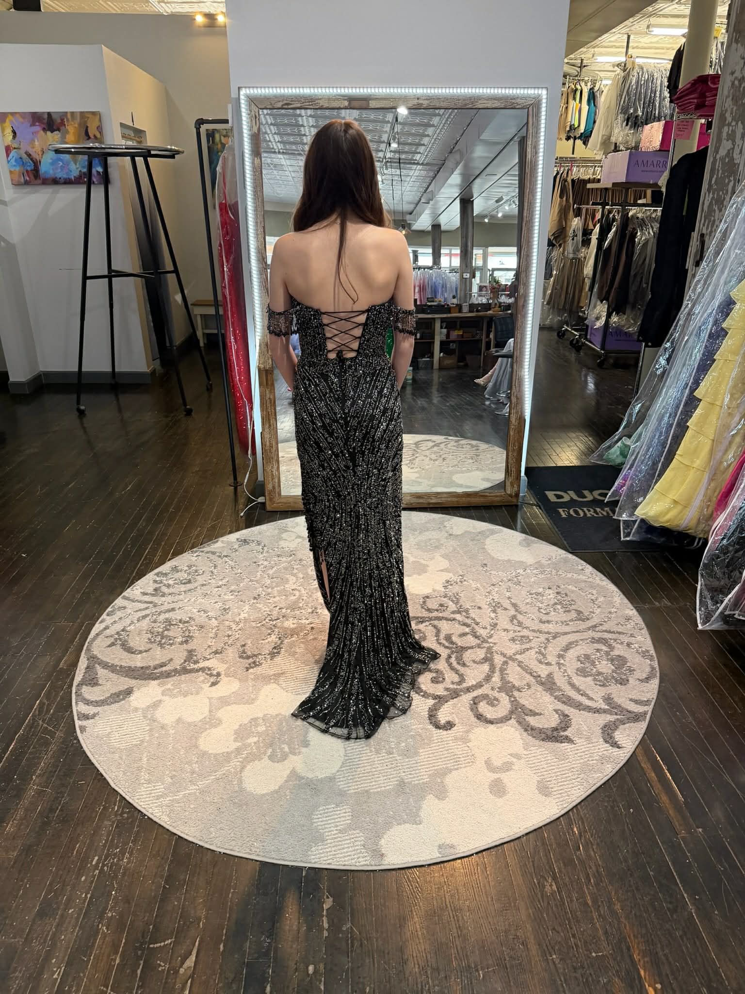 A person stands in a dress shop, facing a mirror and wearing a vibrant, floor-length orange satin gown with a laced back.
