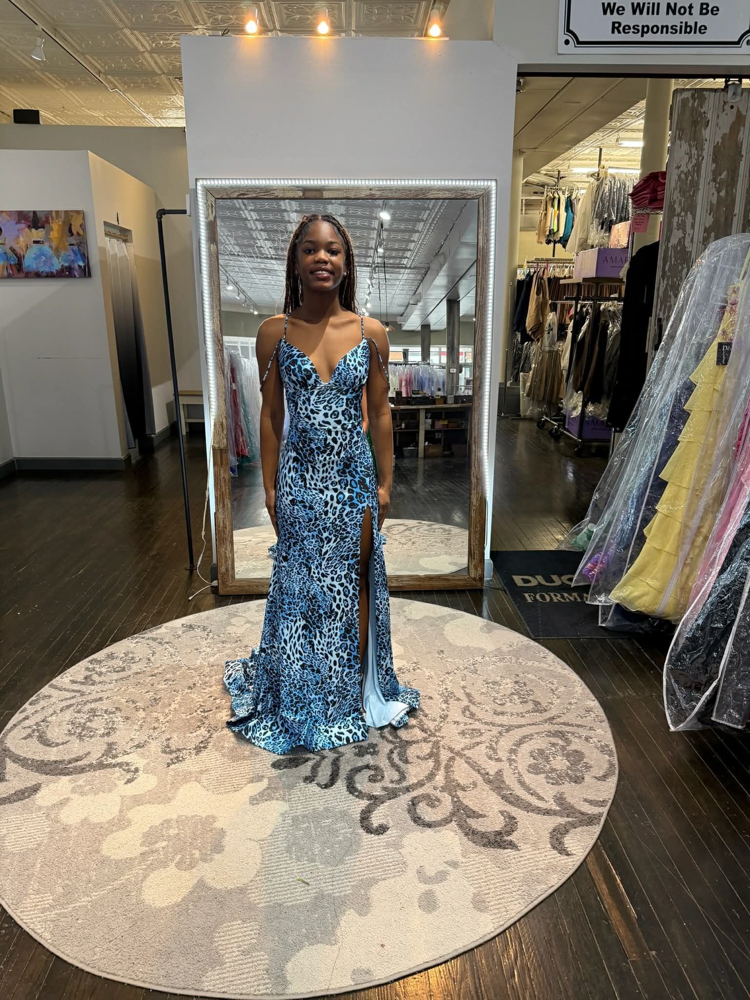 A person stands on a circular rug, facing a large mirror, modeling a floor-length blue and white patterned formal gown.