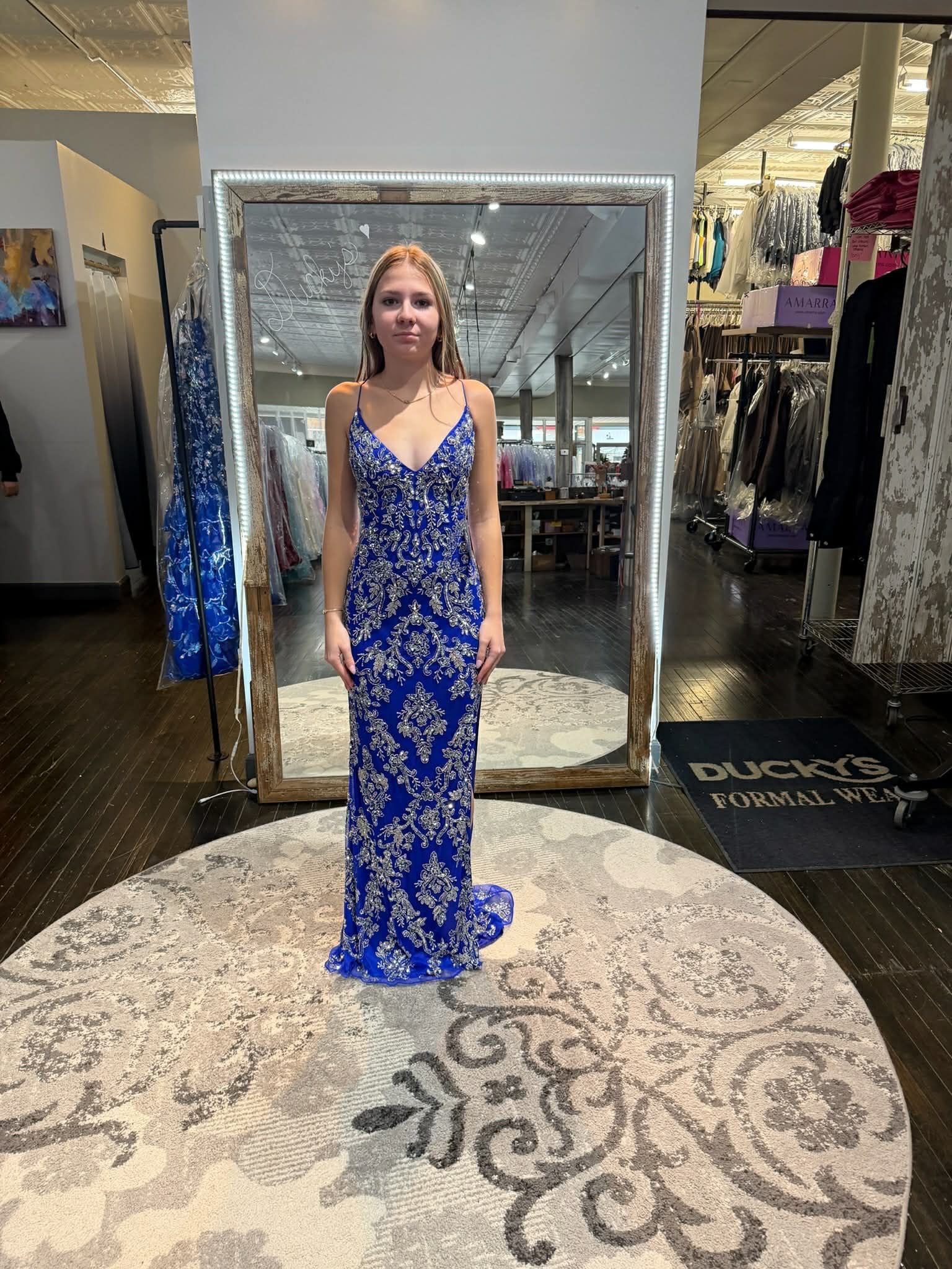 A smiling person wears an off-the-shoulder, floor-length dark green sequined gown in a clothing store with mirrors.
