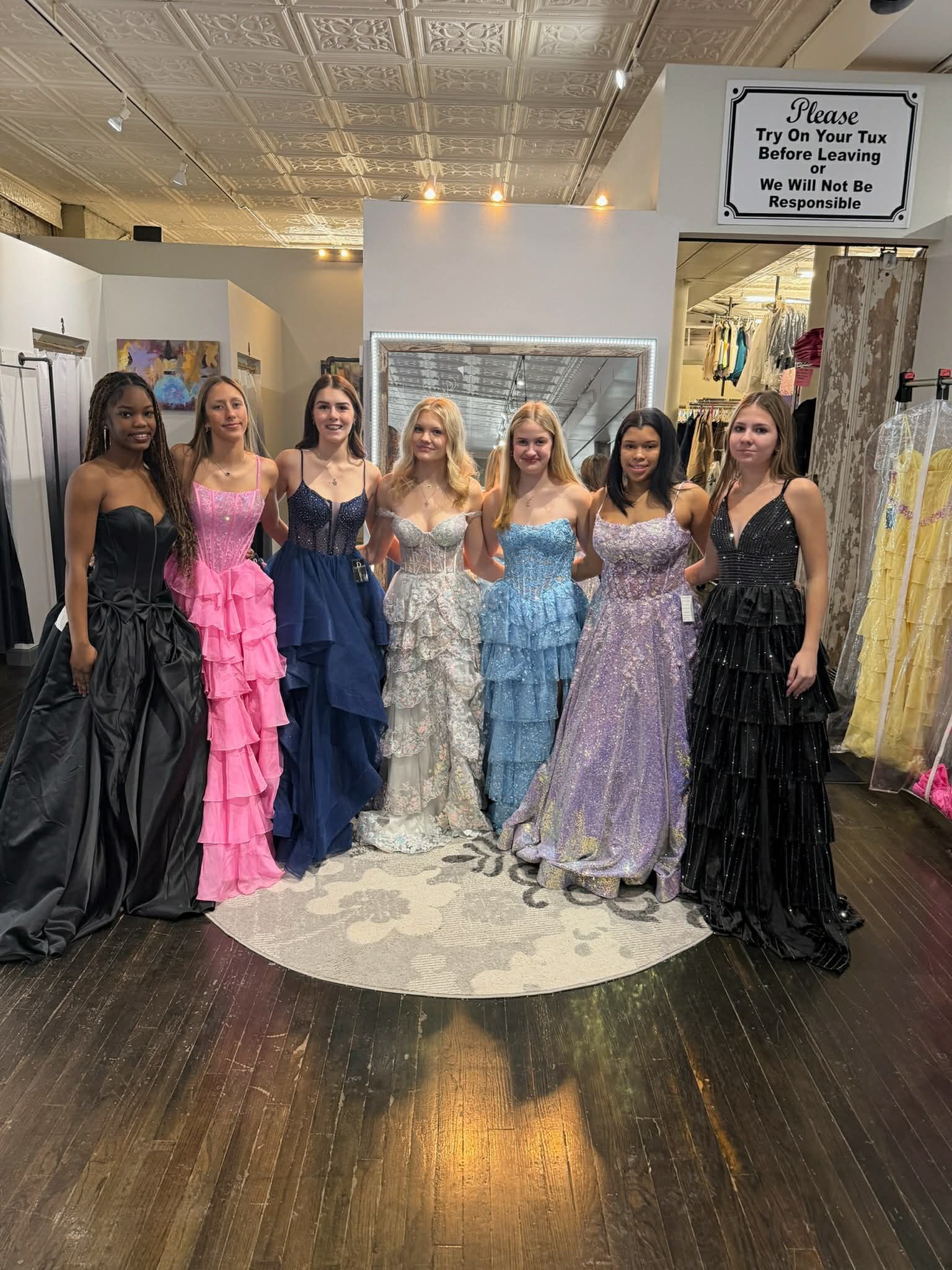 A person wearing a bright yellow, sequined, high-low gown stands on a round patterned rug in a clothing store.