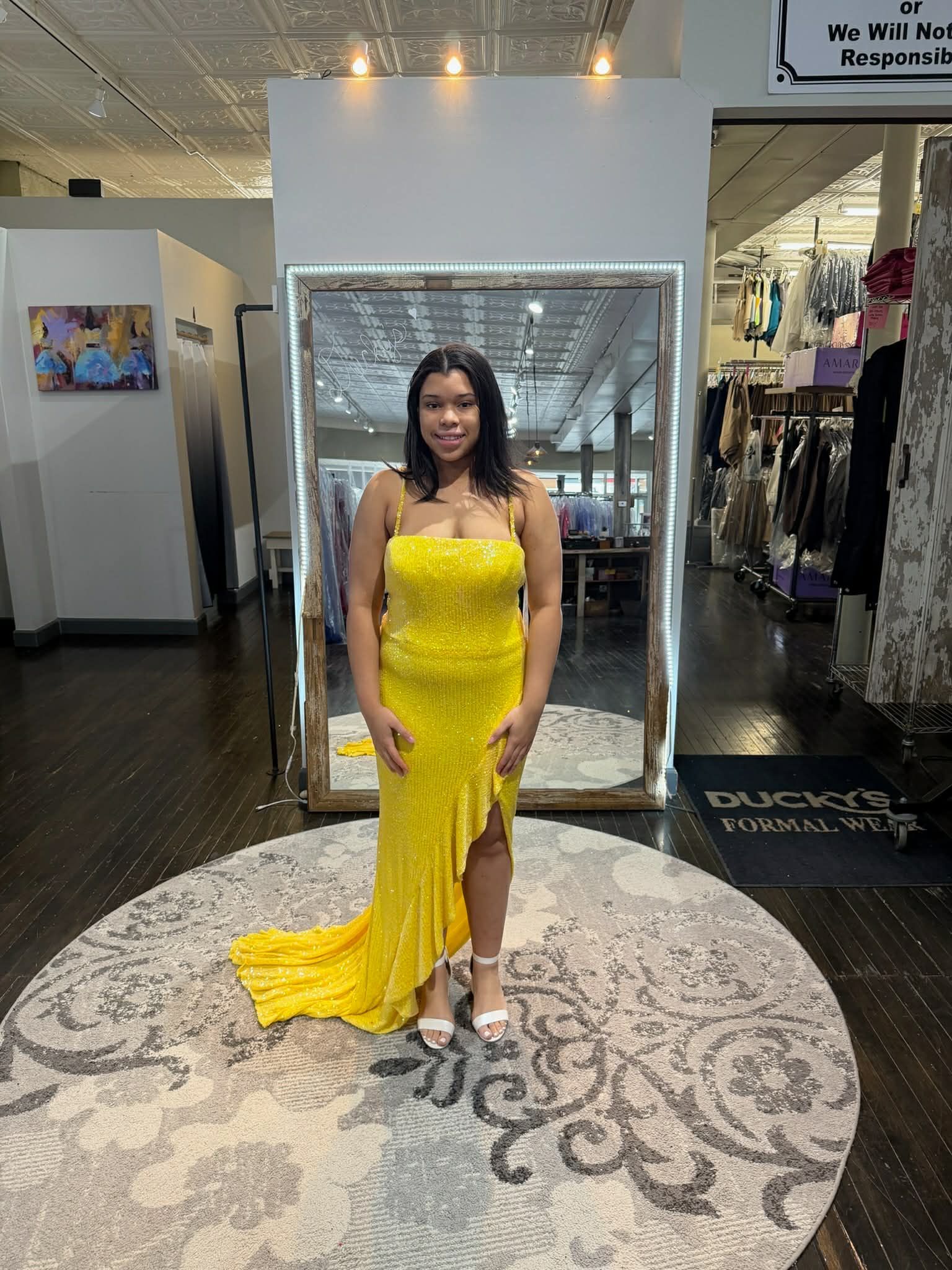 A person stands in a clothing store mirror wearing a light-colored, off-the-shoulder, tiered lace floor-length gown.