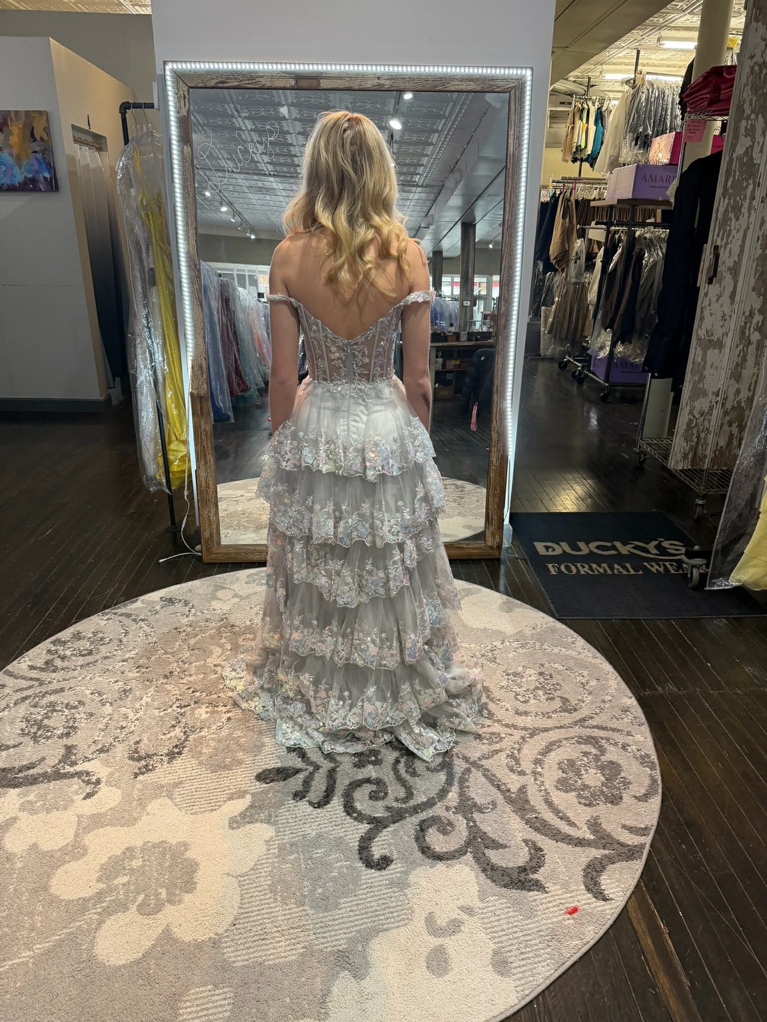 A person stands in a dress shop, facing a mirror and wearing a floor-length, bright orange corset-back evening gown.