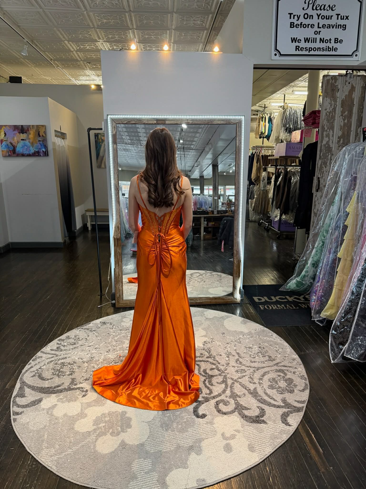 A person wearing a magenta evening gown stands on a circular rug in a clothing store, facing a mirror.