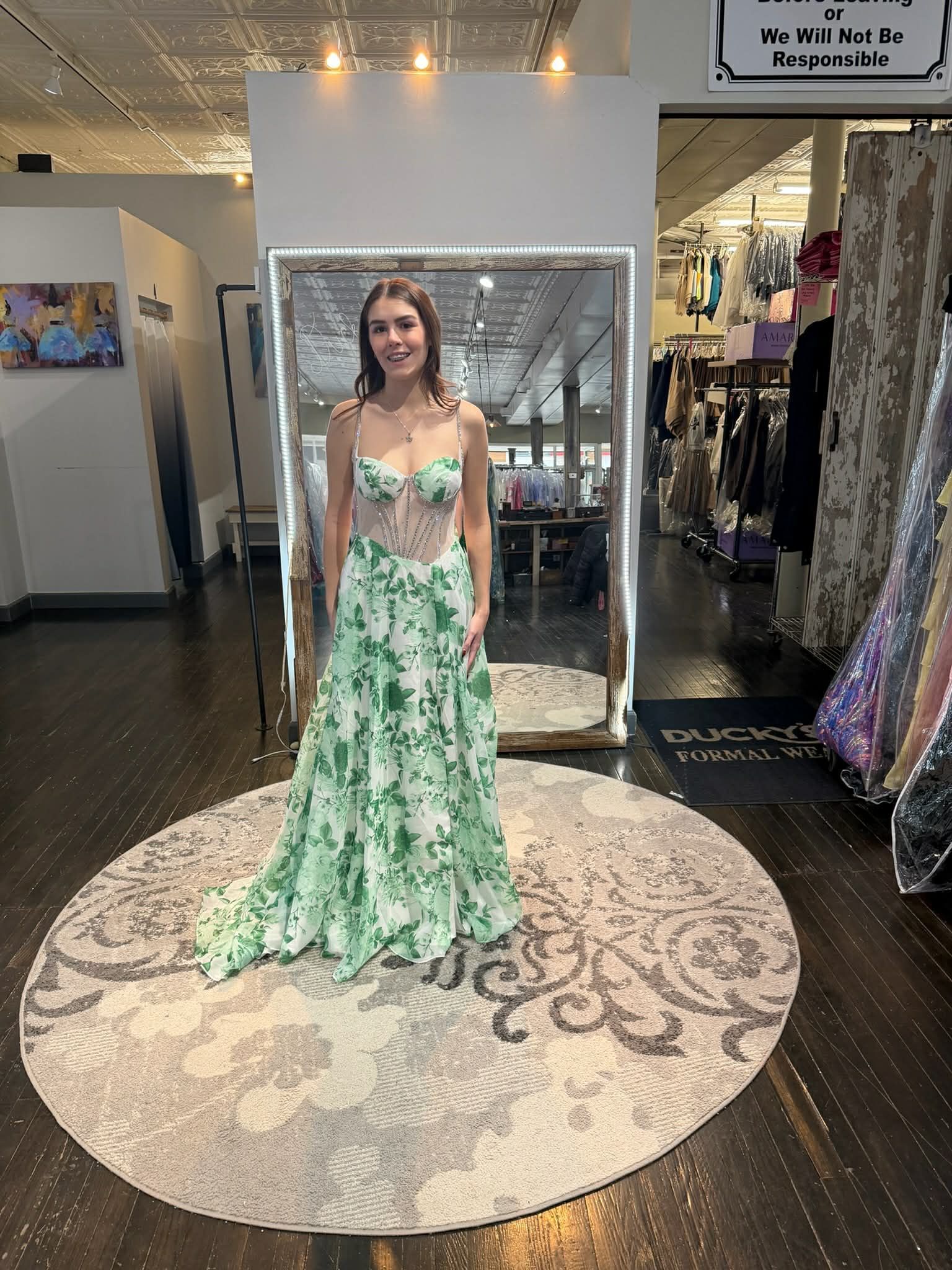Two people stand back-to-back in a dress shop, wearing a green sequined gown and a tiered pink off-the-shoulder gown.