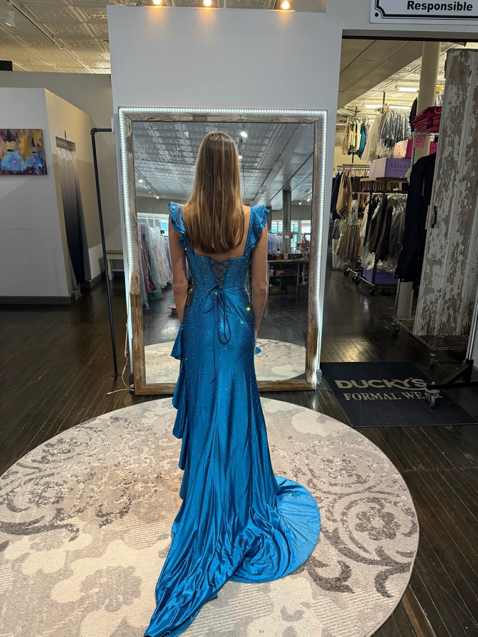 Three people stand in a clothing store, seen from behind, wearing blue, black, and purple formal floor-length gowns.