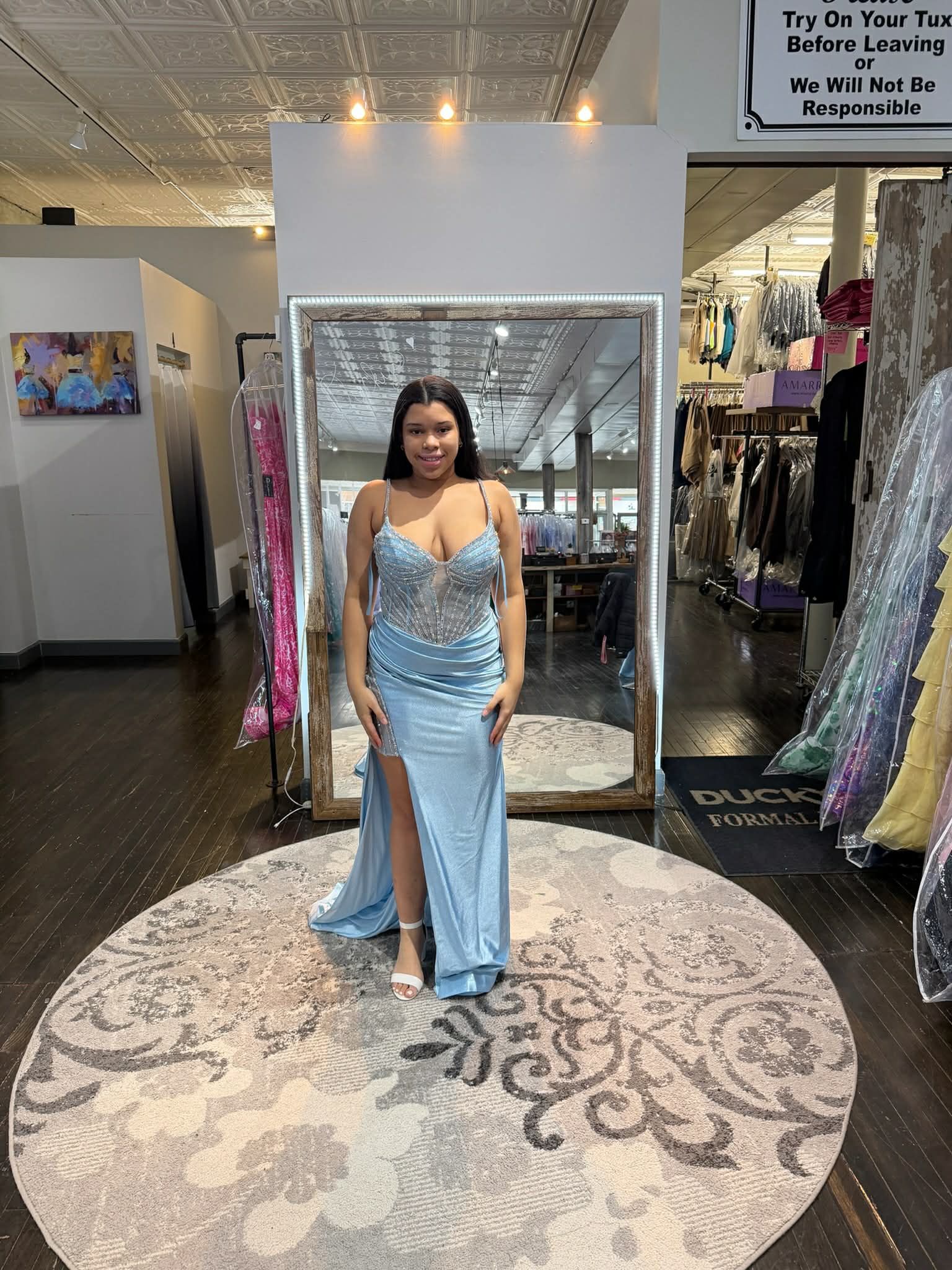 A person stands on a round rug in a boutique, wearing a floor-length, off-the-shoulder, black sequined gown in front of a mirror.