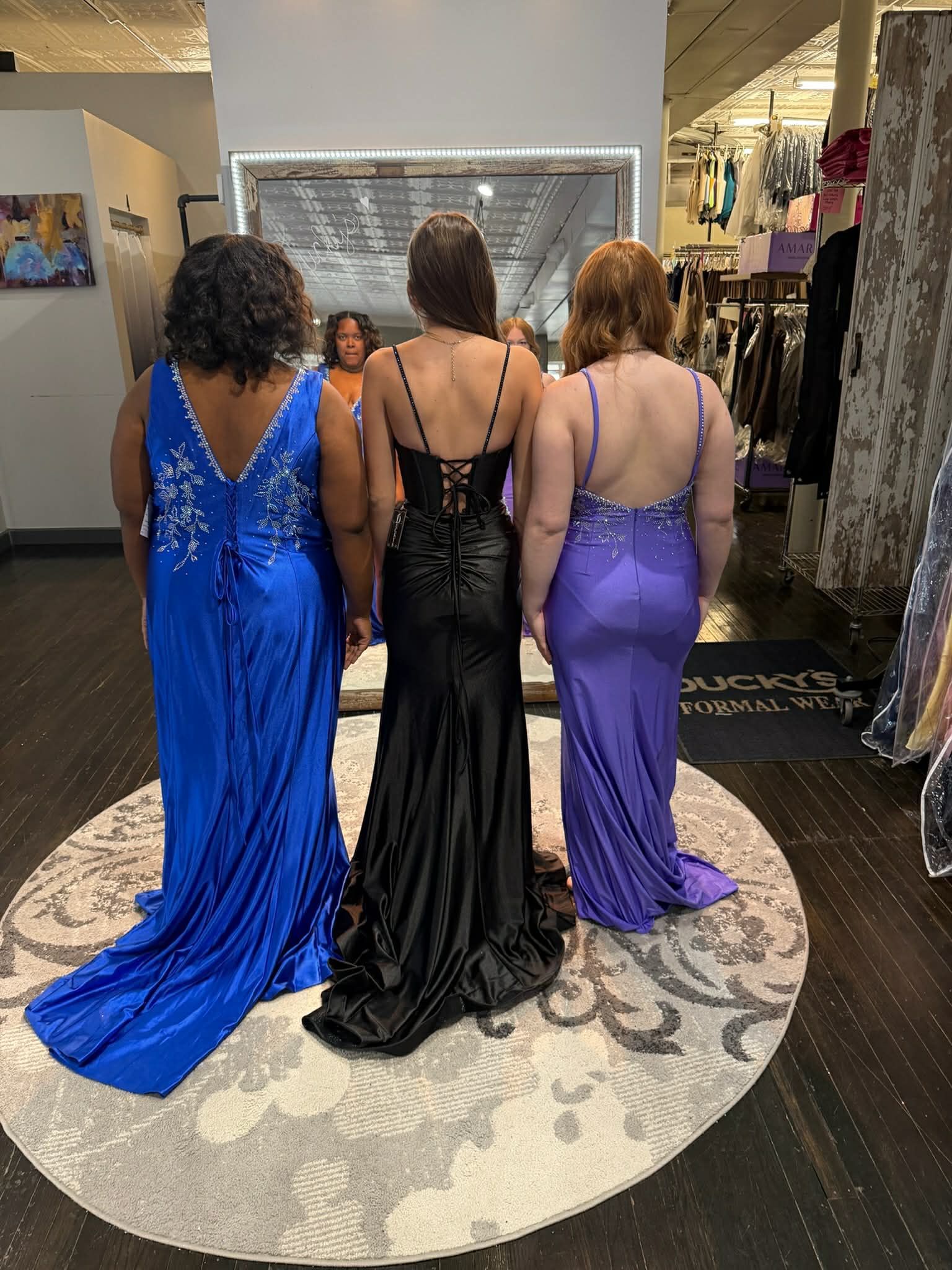 A person with red hair stands in front of a mirror wearing a floor-length lavender dress with a ruffled skirt and back.