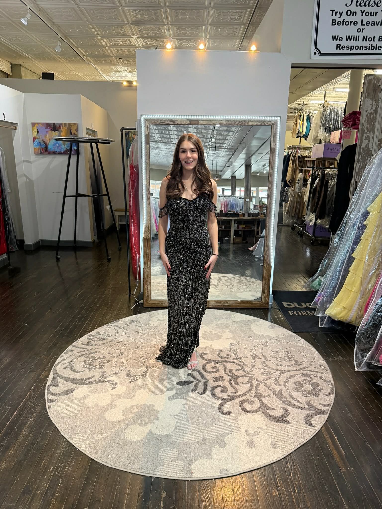 A person stands in a clothing boutique, facing a large mirror while wearing a floor-length, coral, sequined backless dress.