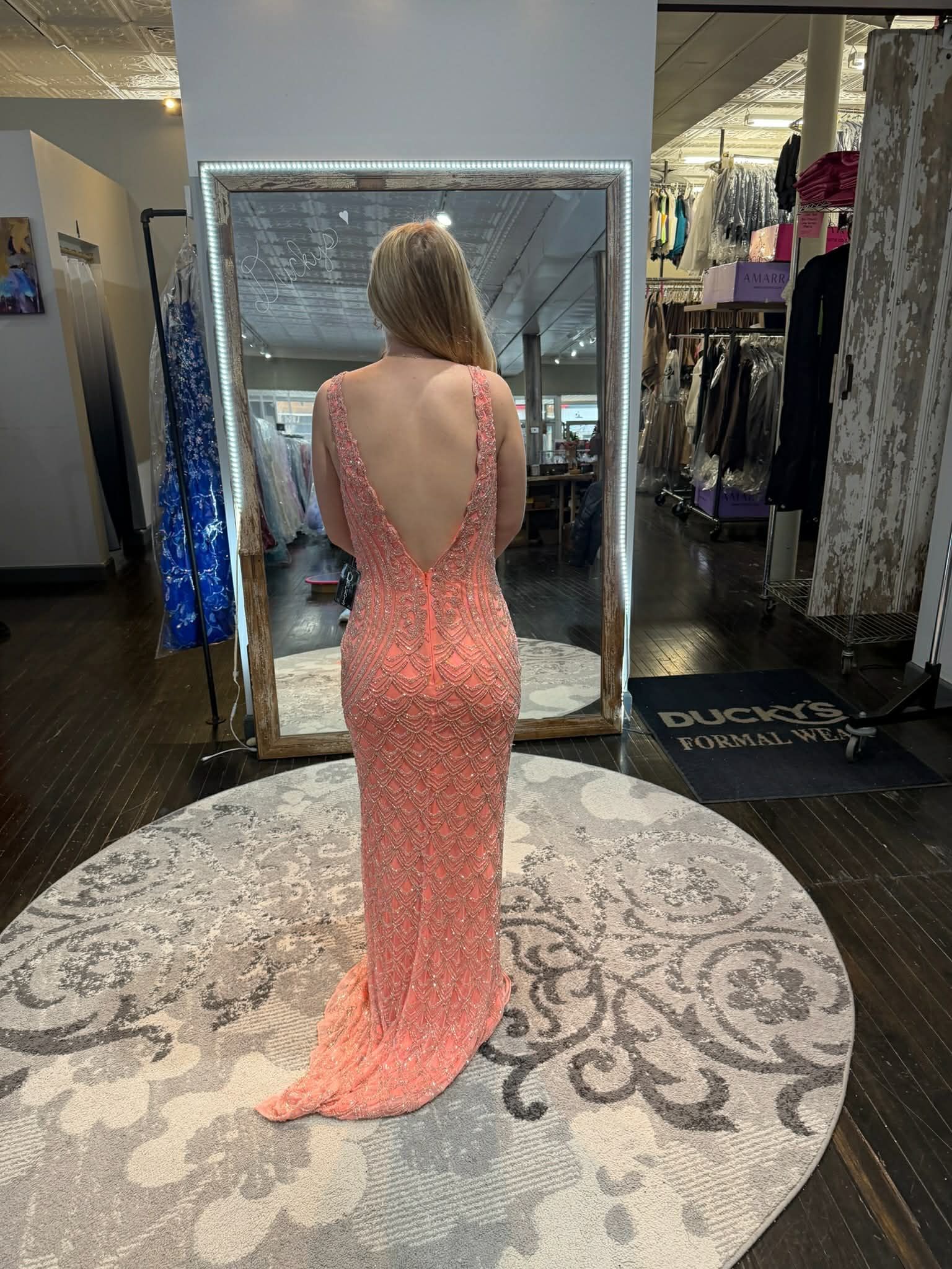 A person stands in a dress shop wearing a black, floor-length, spaghetti-strap gown on a large, circular patterned rug.