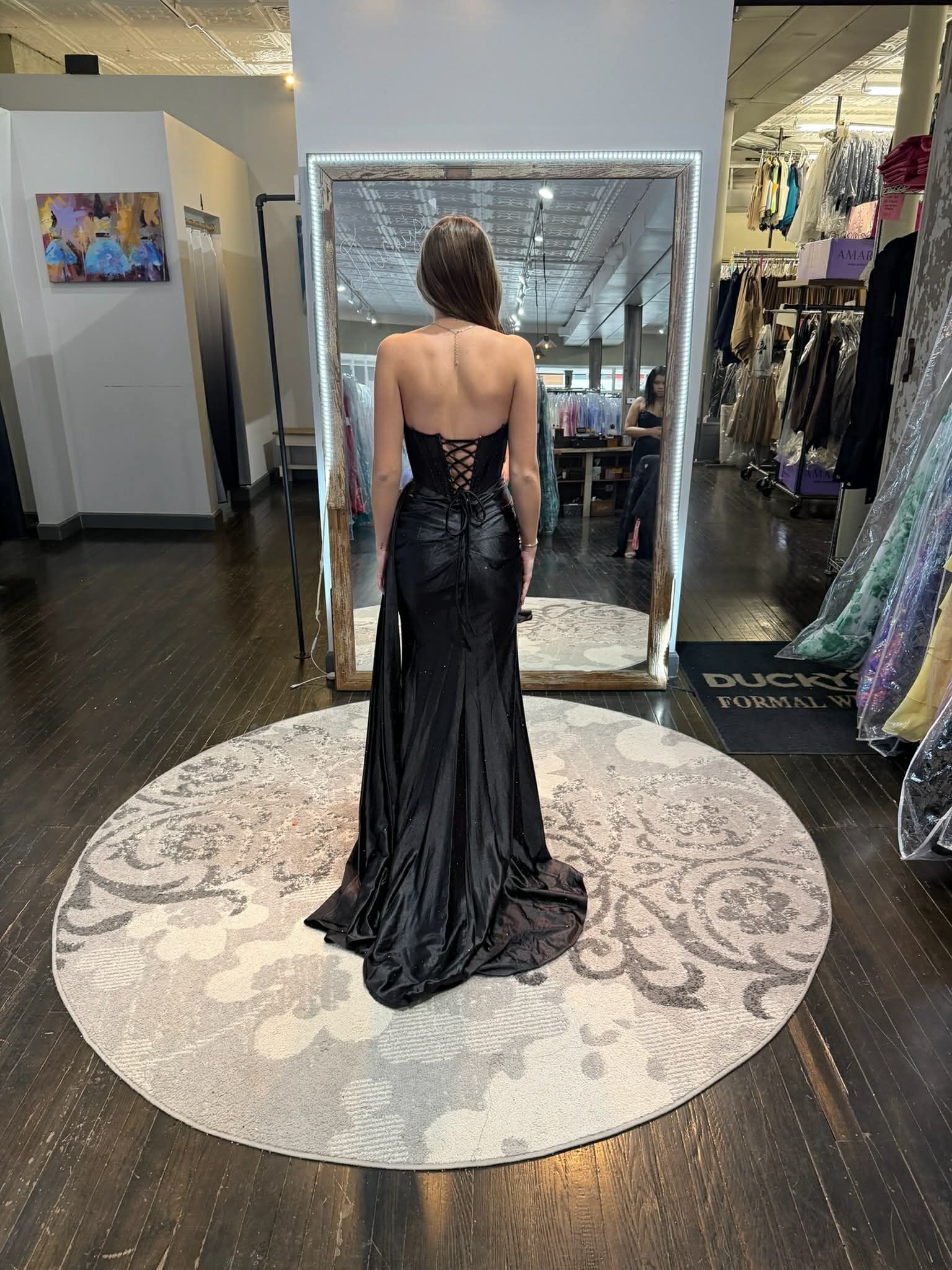 A person wears a floor-length, bright orange mermaid-style gown with a lace-up back while standing in a clothing boutique.