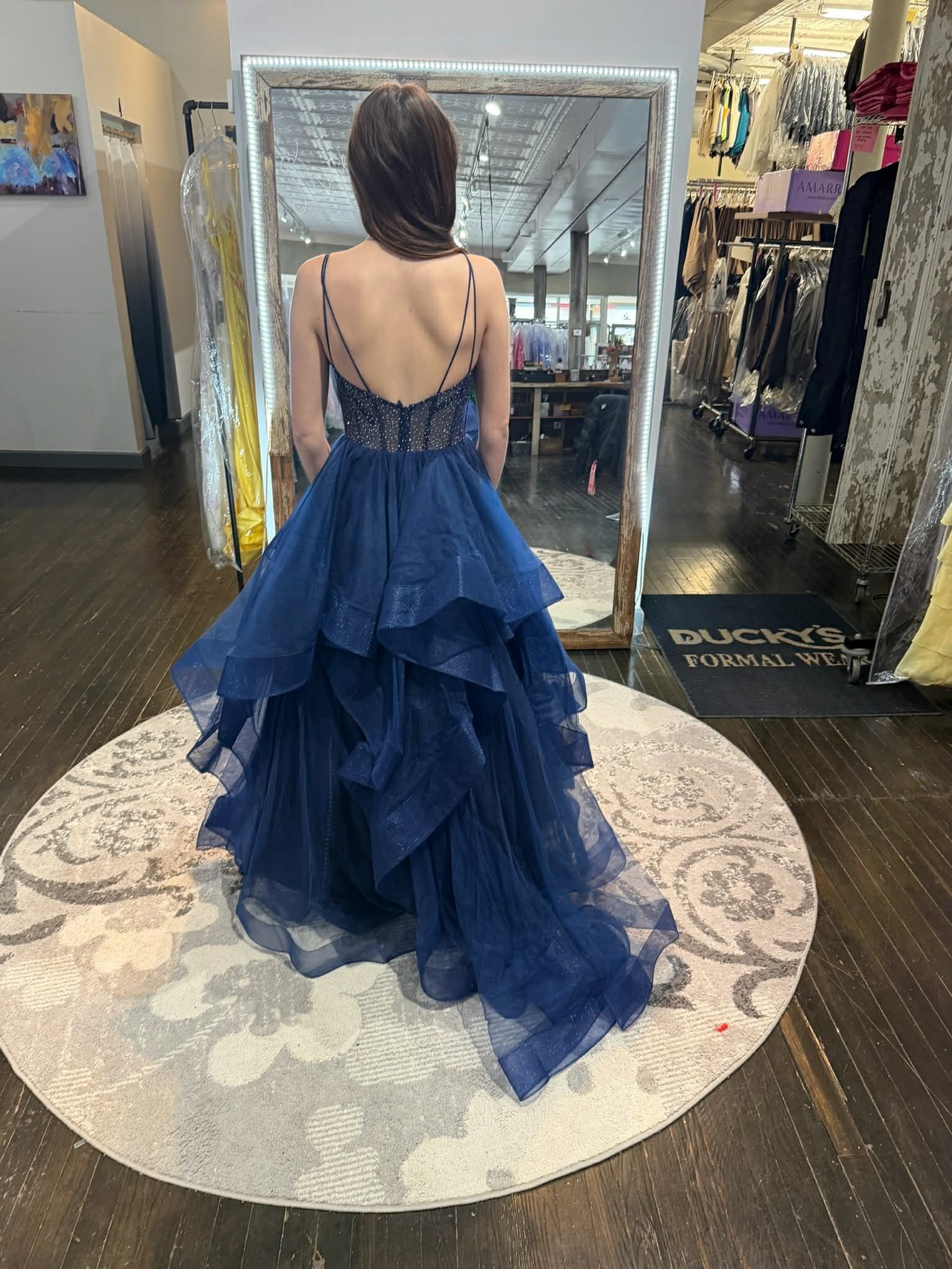 A person stands in a clothing store, facing a mirror, wearing a long, vibrant purple and blue sequined gown with a lace-up back.