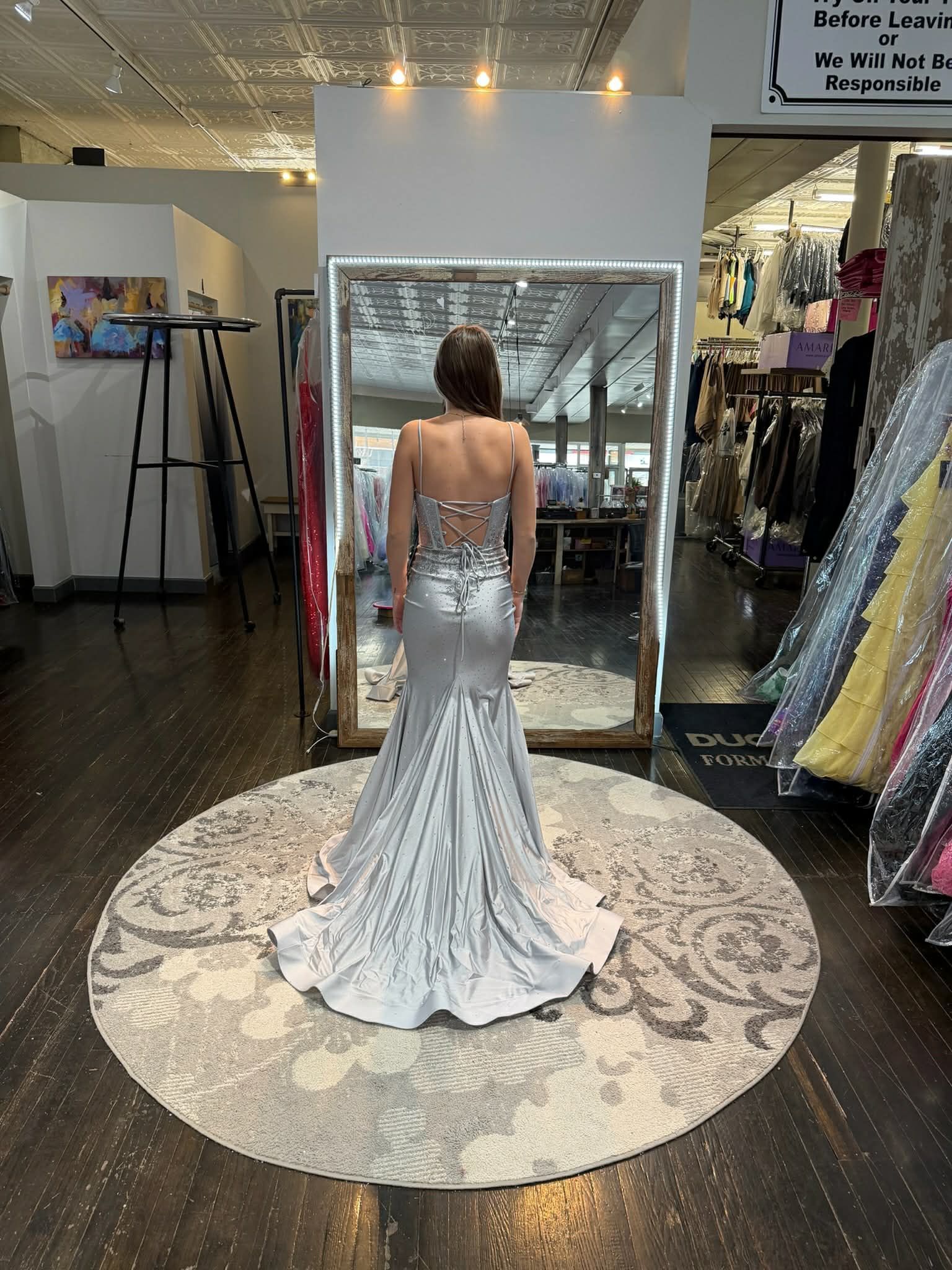A person stands facing a large mirror in a boutique, wearing a backless, floor-length silver sequined mermaid-style gown.