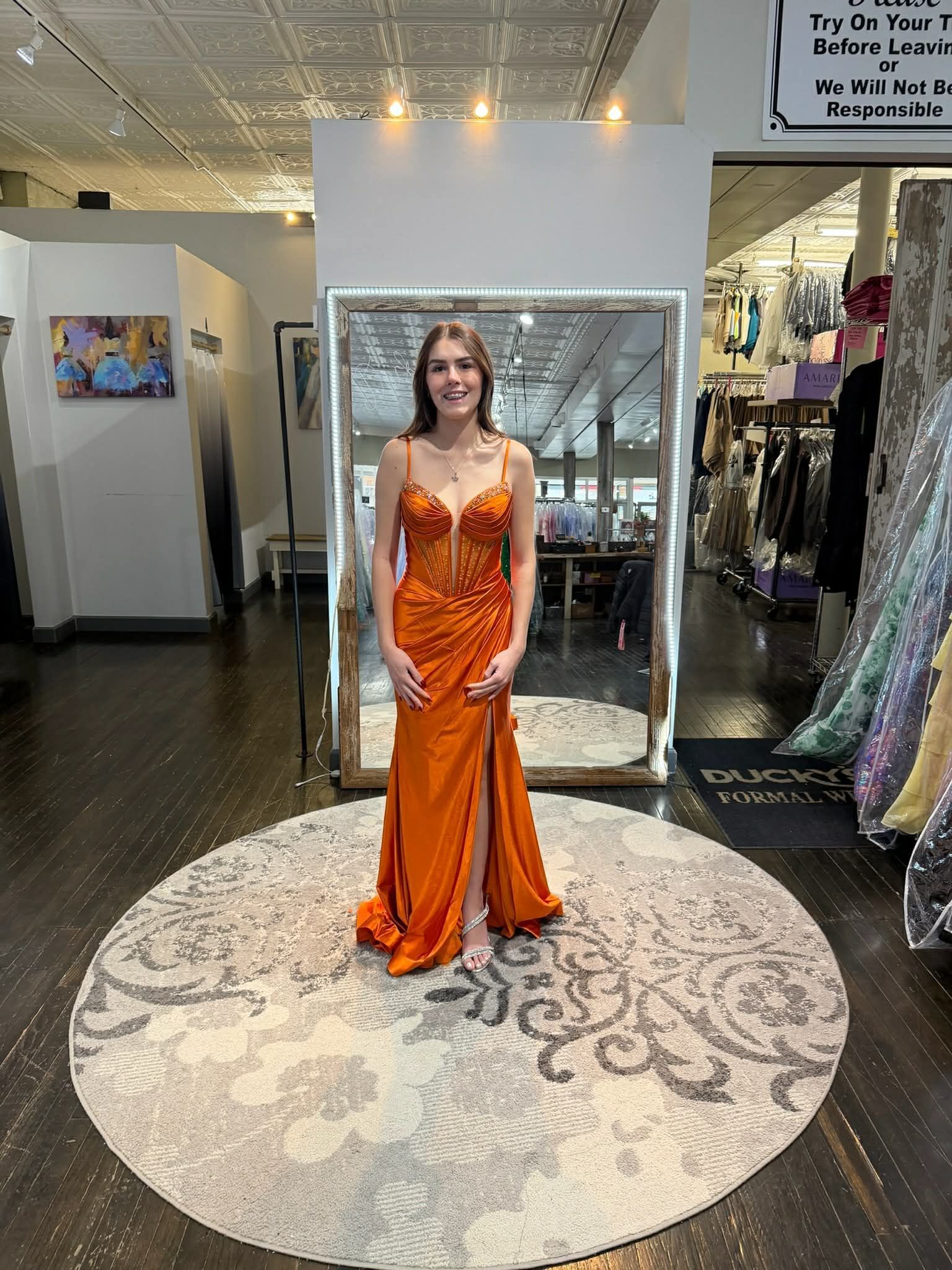 A person smiling while wearing a long, sleeveless, rust-orange gown with a high slit, standing in a formal wear boutique.