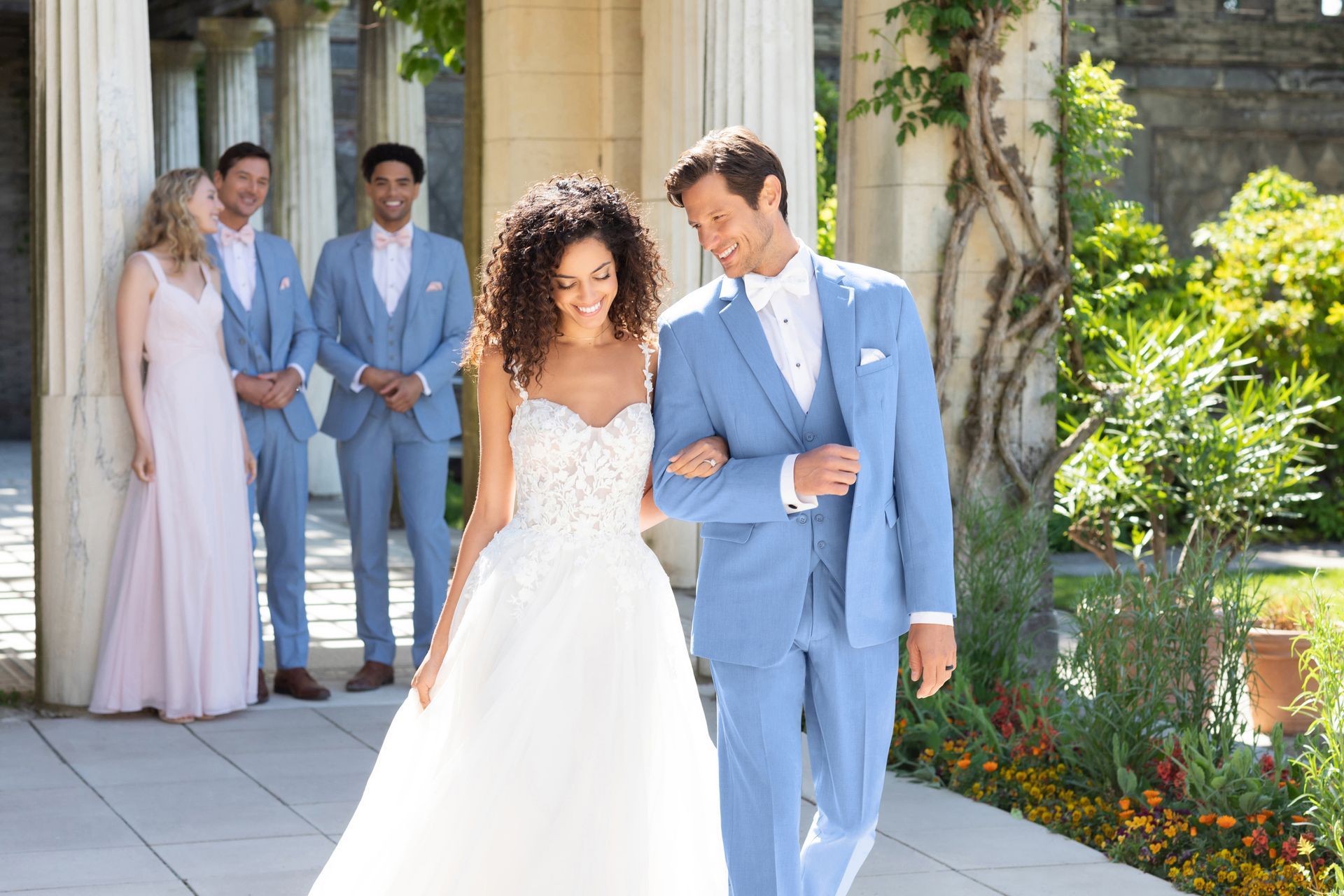 A bride and groom in a light blue suit walk arm-in-arm in a garden, with two others standing in the background.