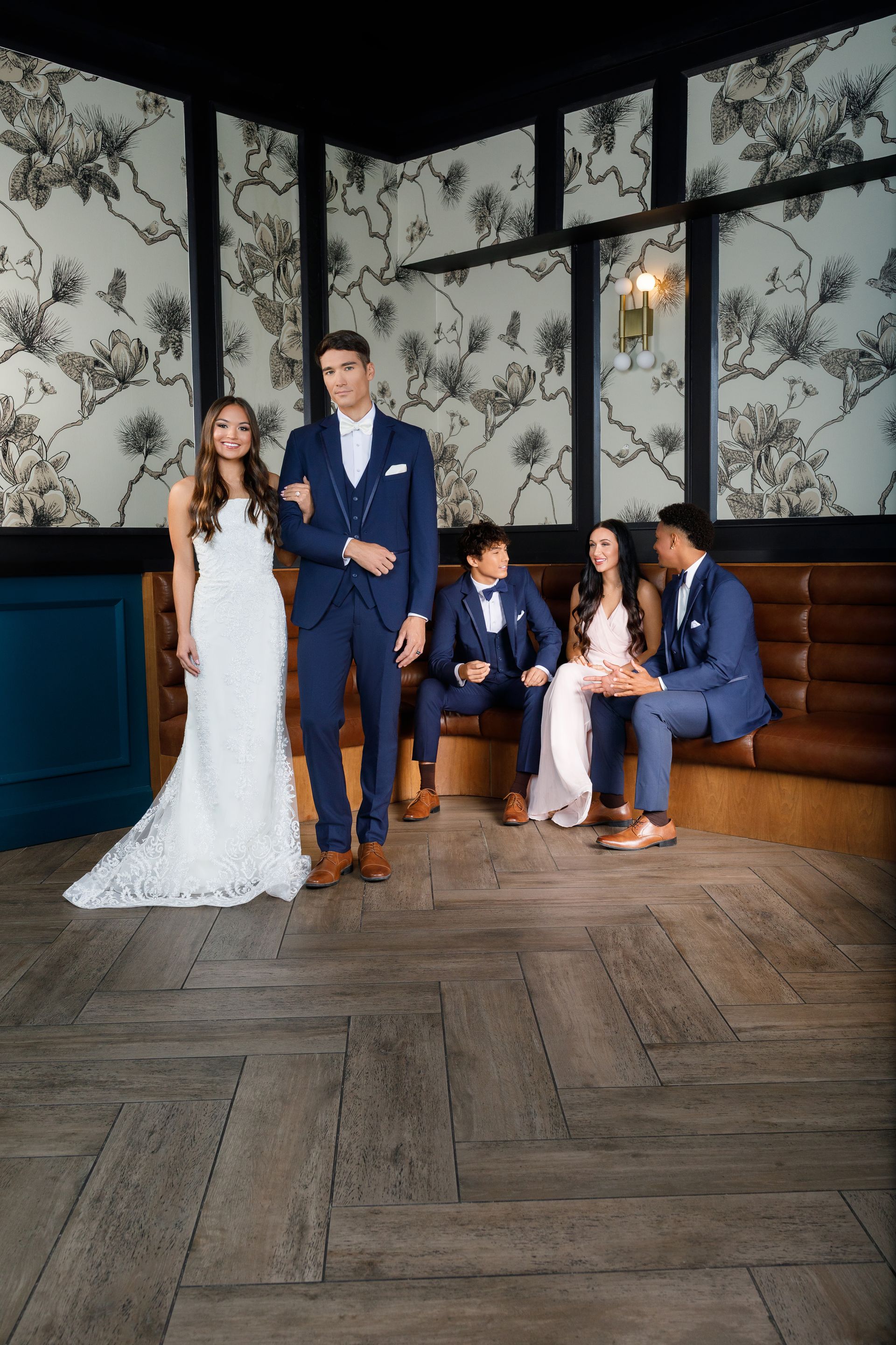 A bride and groom stand in the foreground, while two guests sit on a leather bench against a floral-patterned wall.