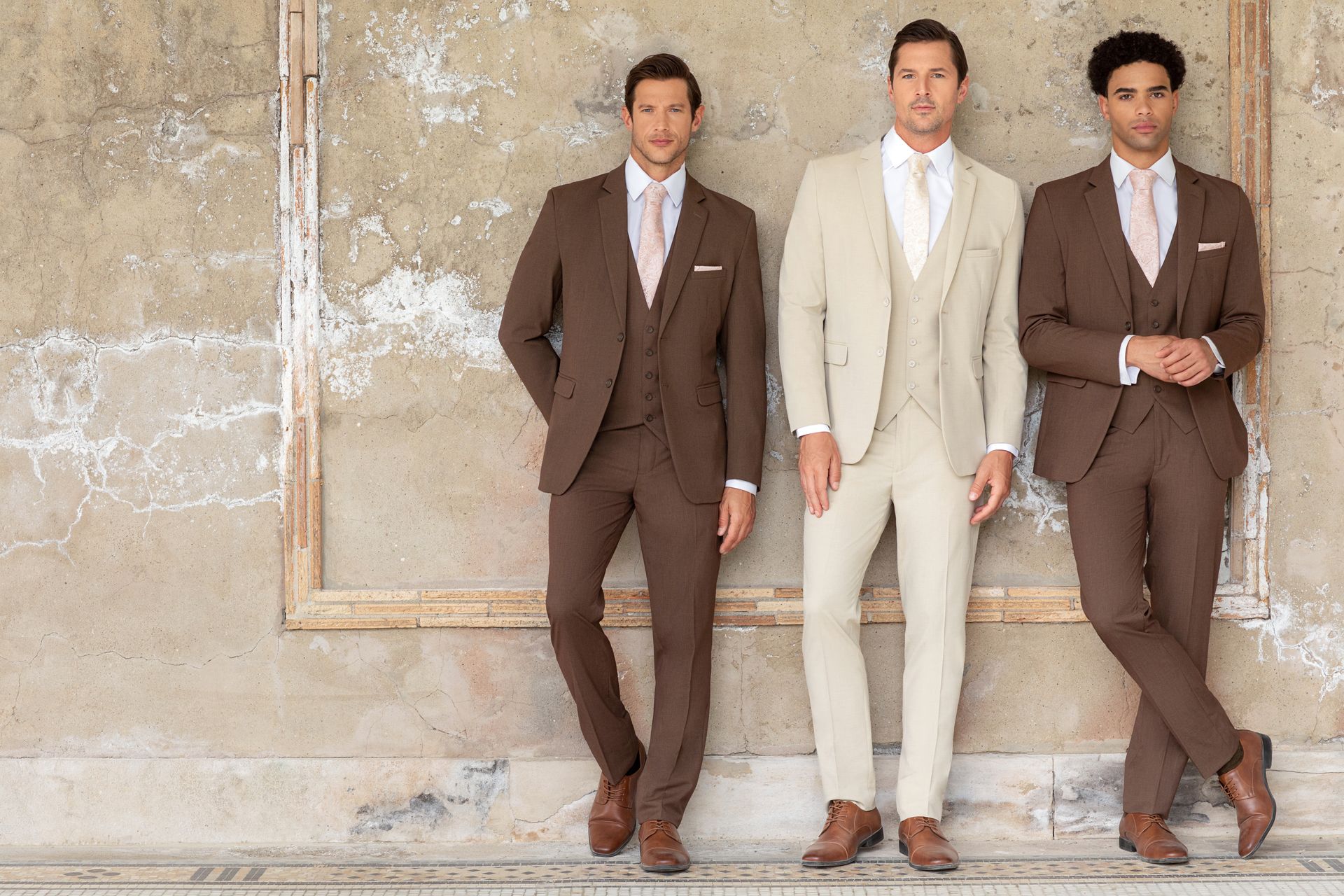 Three models posing against a textured concrete wall, wearing brown and cream three-piece suits with ties.