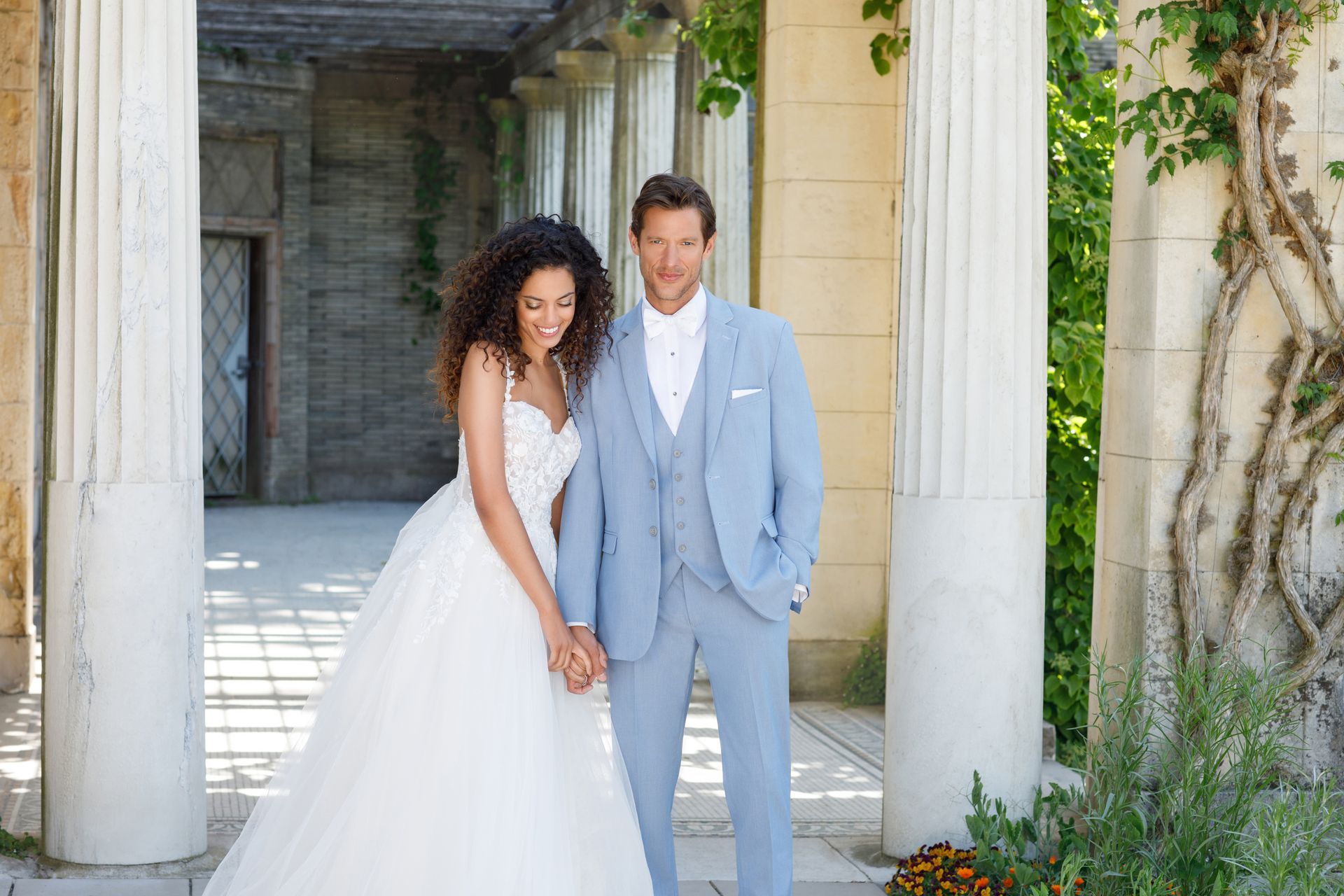 A couple standing together in a sunlit stone colonnade, wearing a formal white wedding dress and a light blue suit.