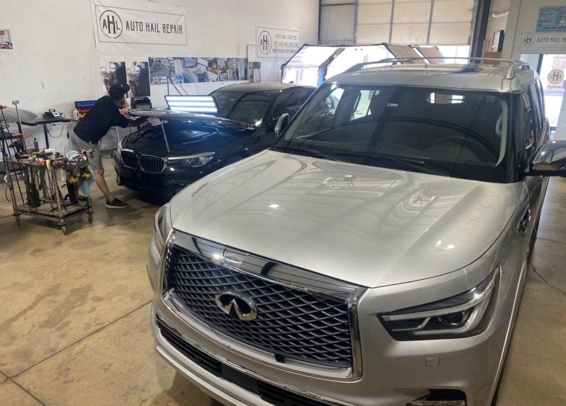 Silver Infiniti SUV in a garage, a person works on a blue car in the background.