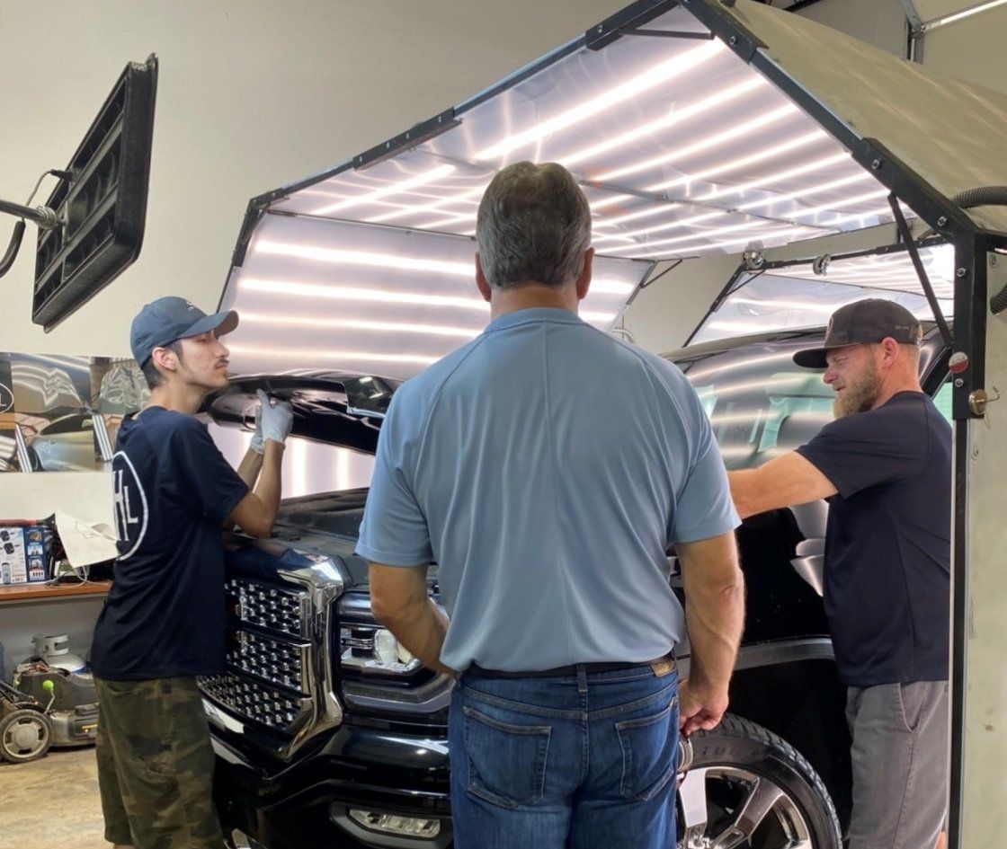 Three men inspect a black truck under a light rig in a garage. Two men work on the hood while the third observes.