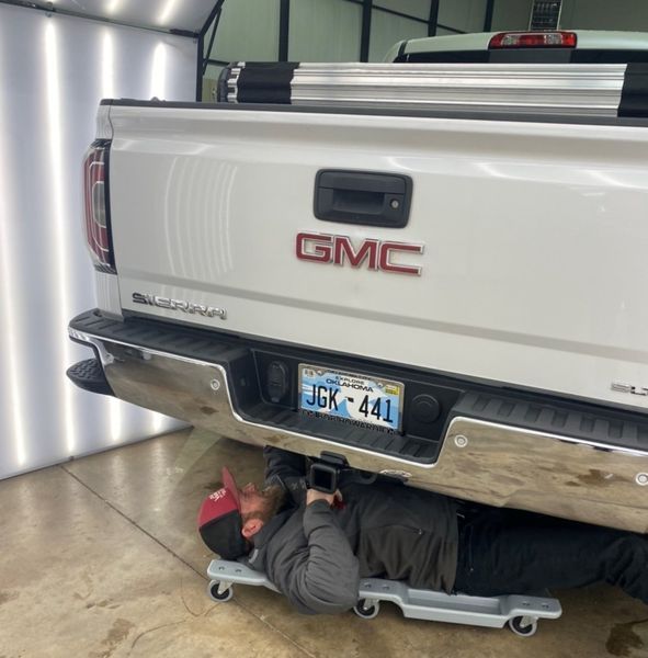 Man working underneath a white GMC Sierra truck, using a rolling cart in a garage.