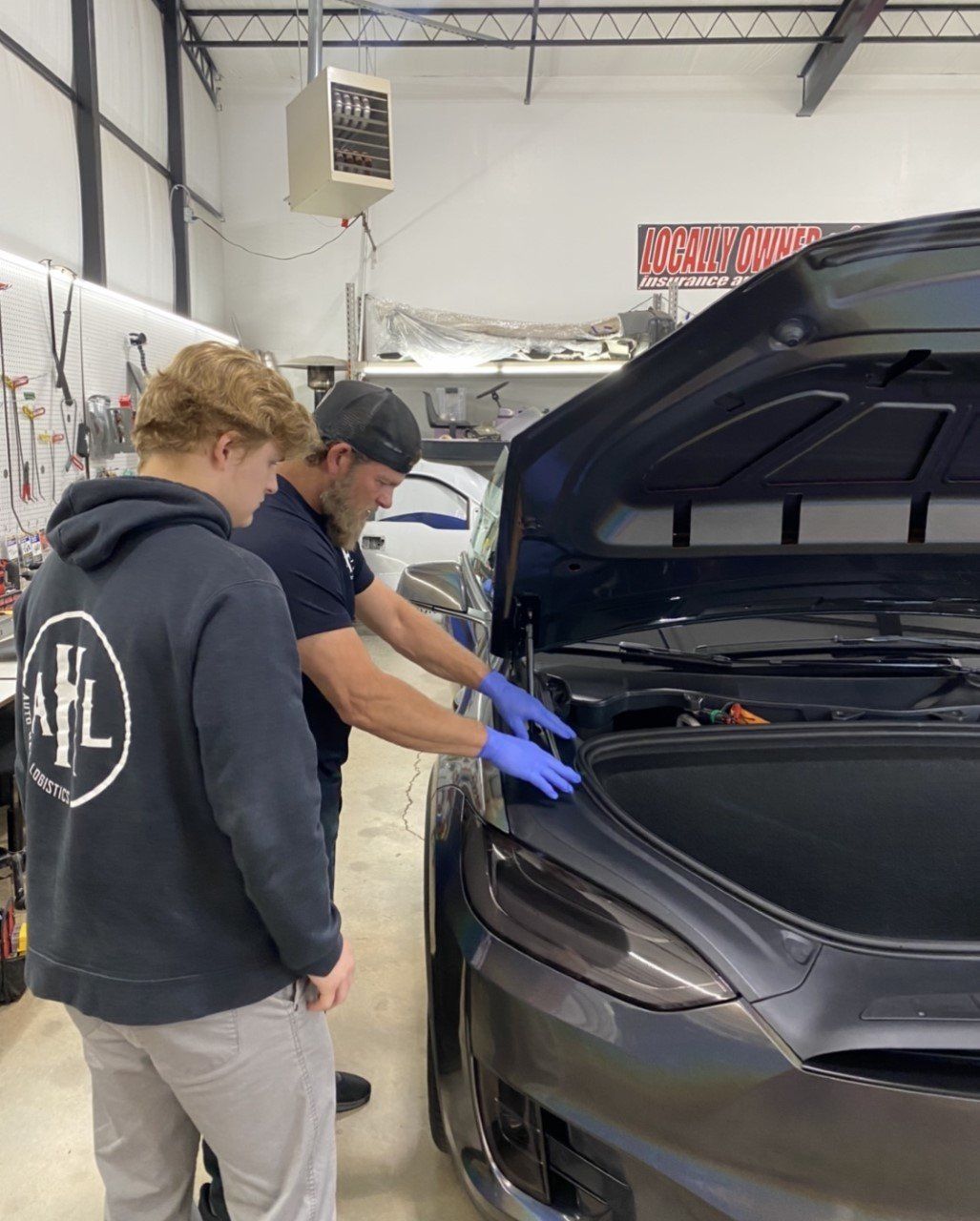 Two people examining a car's open hood in a garage. One wears blue gloves.